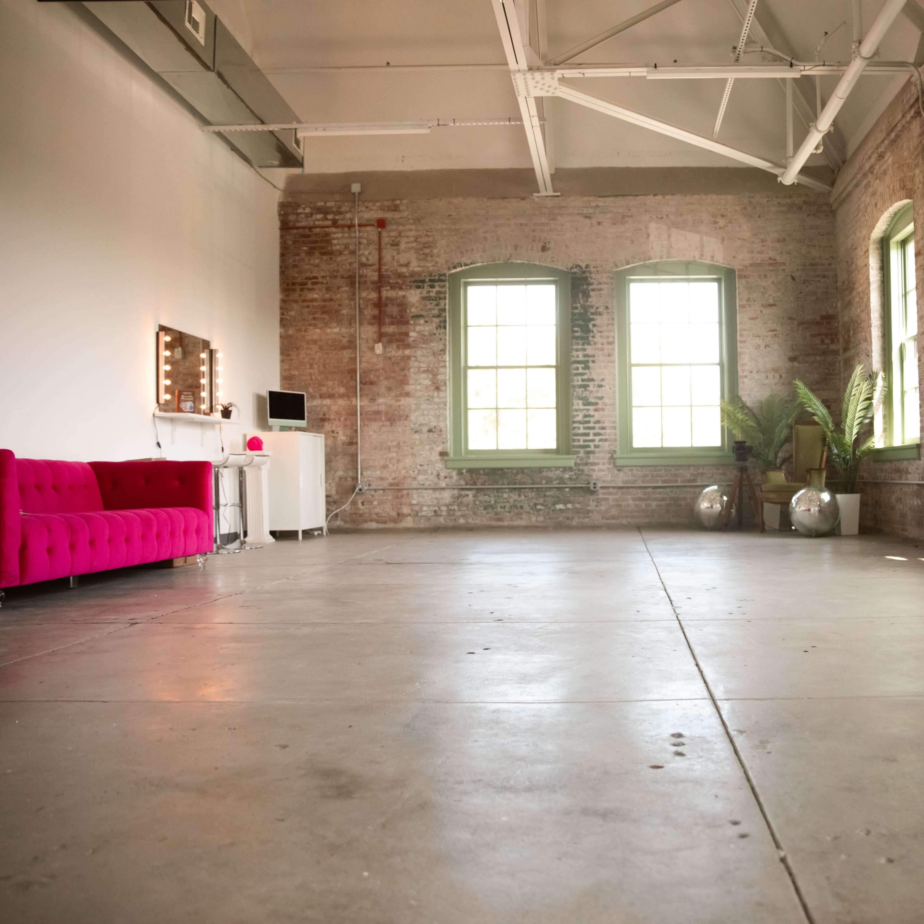 A spacious room with brick walls, two large green-framed windows, a pink couch, a white vanity with a mirror, and two decorative silver spheres.