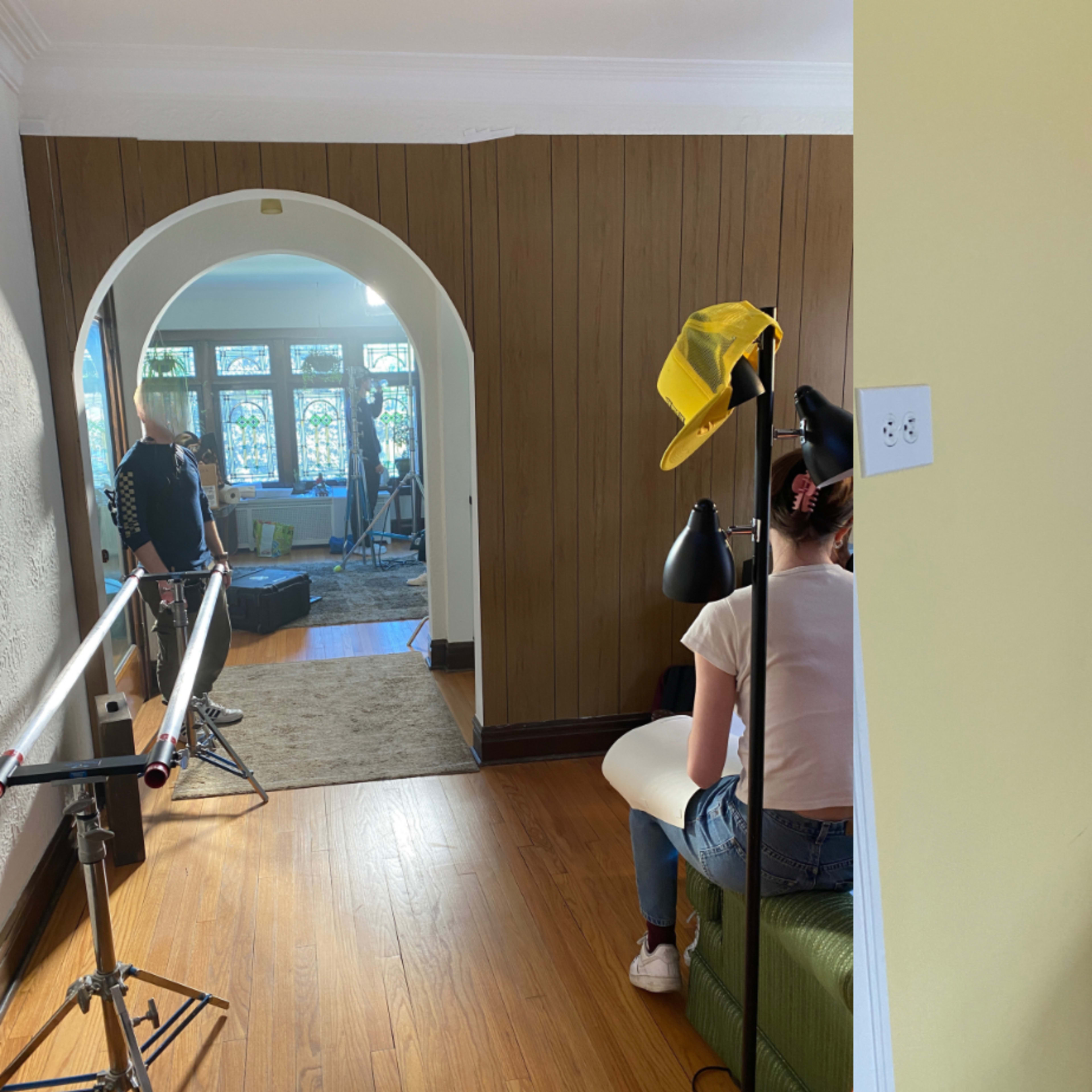 Two sections of a film set: one side features a person sitting near a camera setup in a room, while the other side shows a hallway with a track for filming.