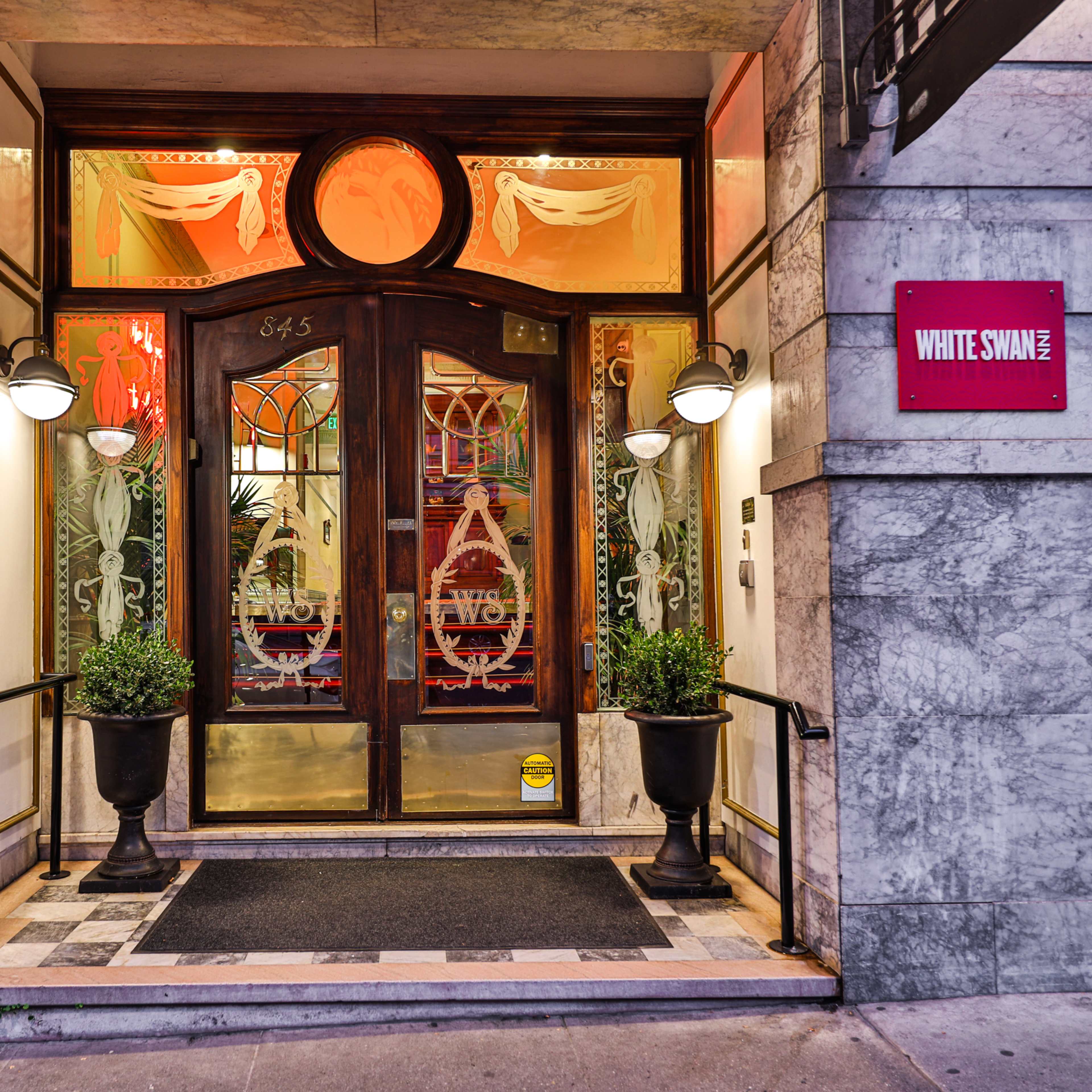 The entrance of the White Swan building features a wooden double door framed by decorative glass panels and potted plants on either side.