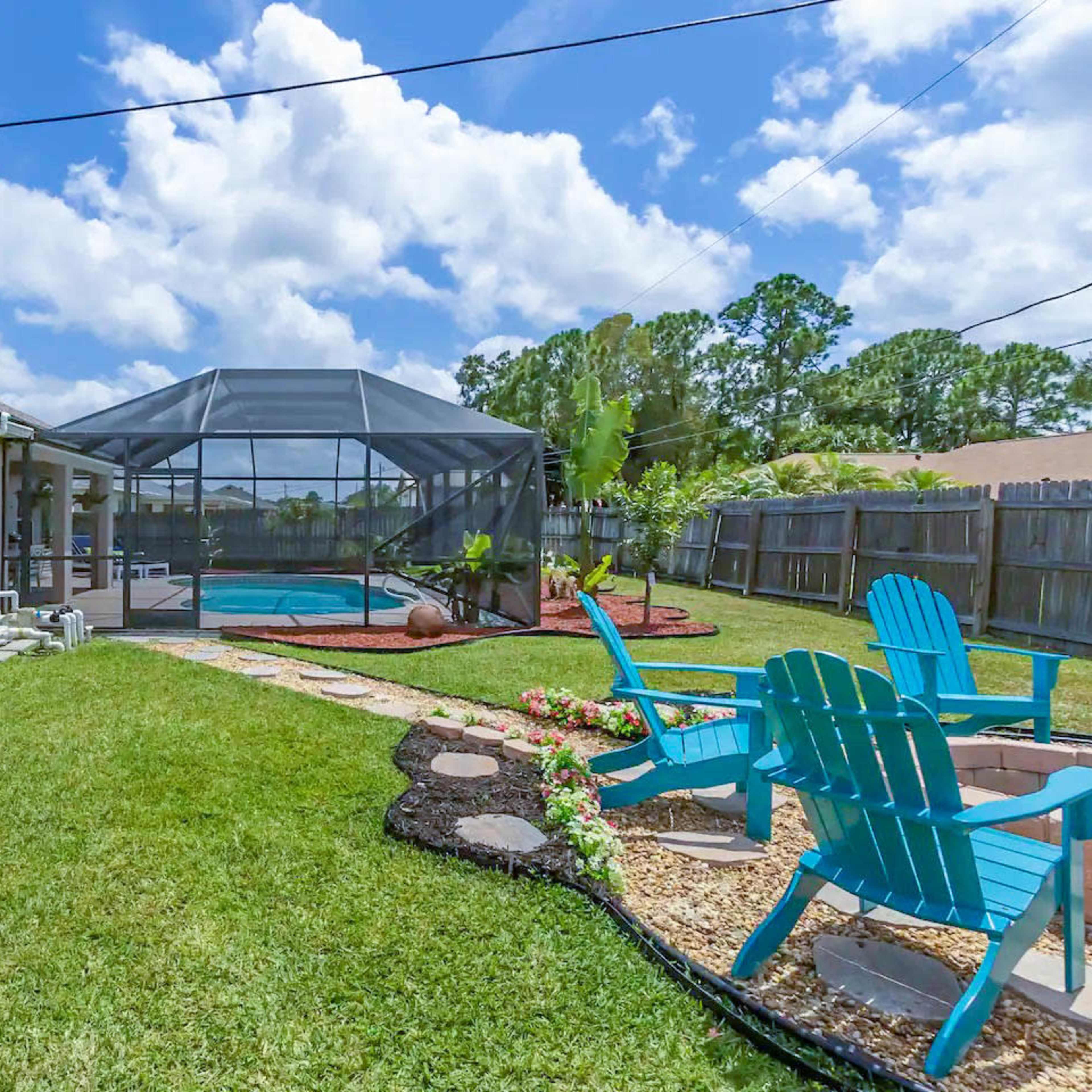 A backyard with a pool enclosed by a screen, surrounded by grass, flower beds, and patio chairs.