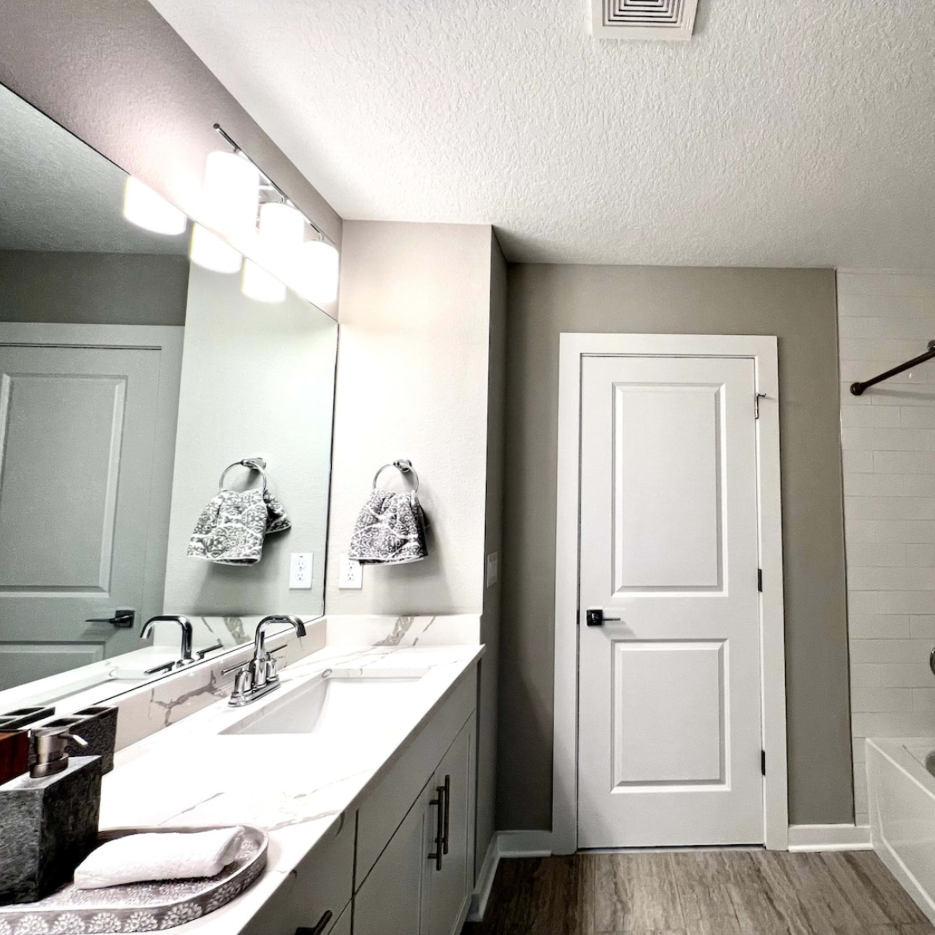 A modern bathroom featuring a bathtub, a wall-mounted mirror, double sinks, and a door leading to another room.