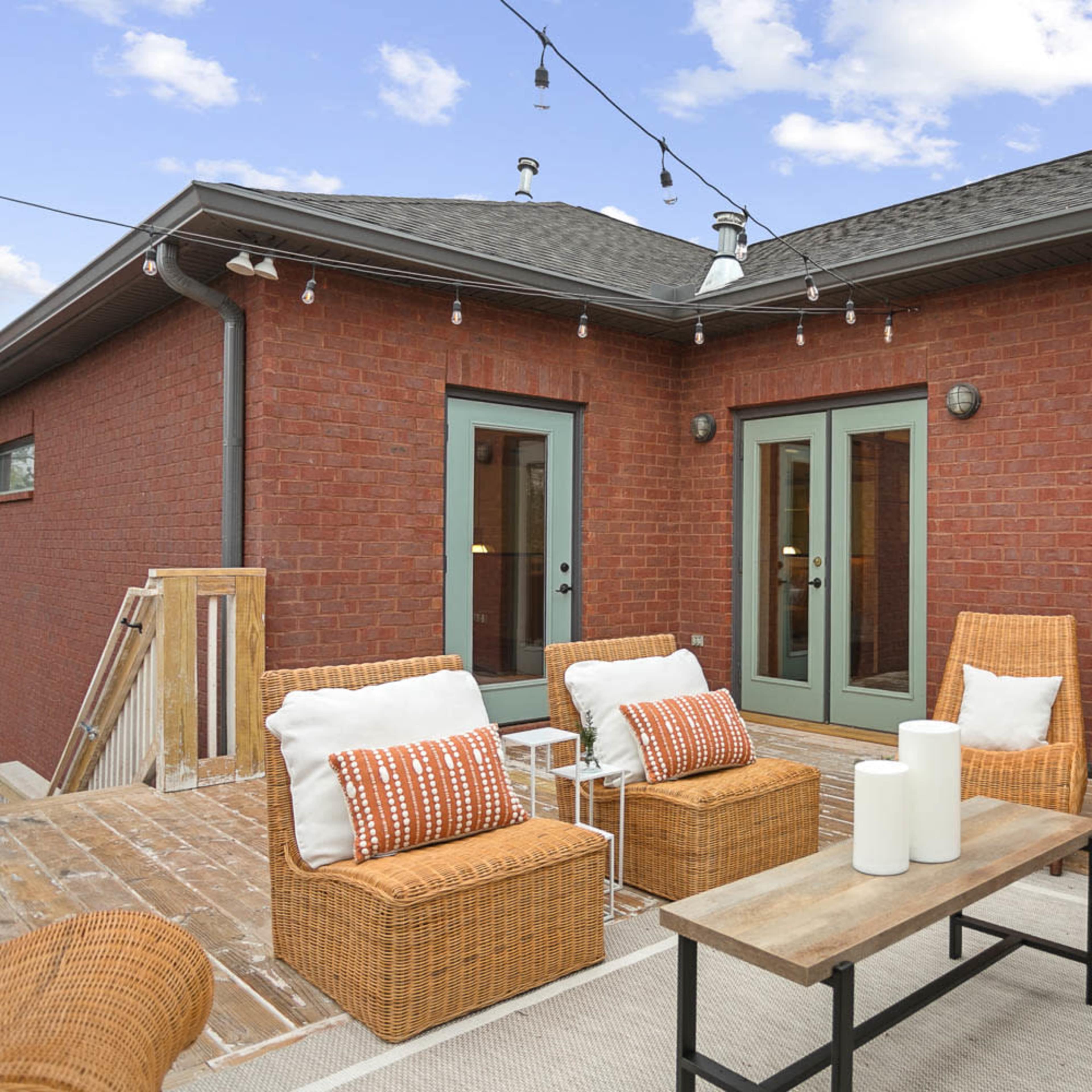 A patio area with wicker chairs, a wooden table, and string lights above, adjacent to a brick wall and glass doors.