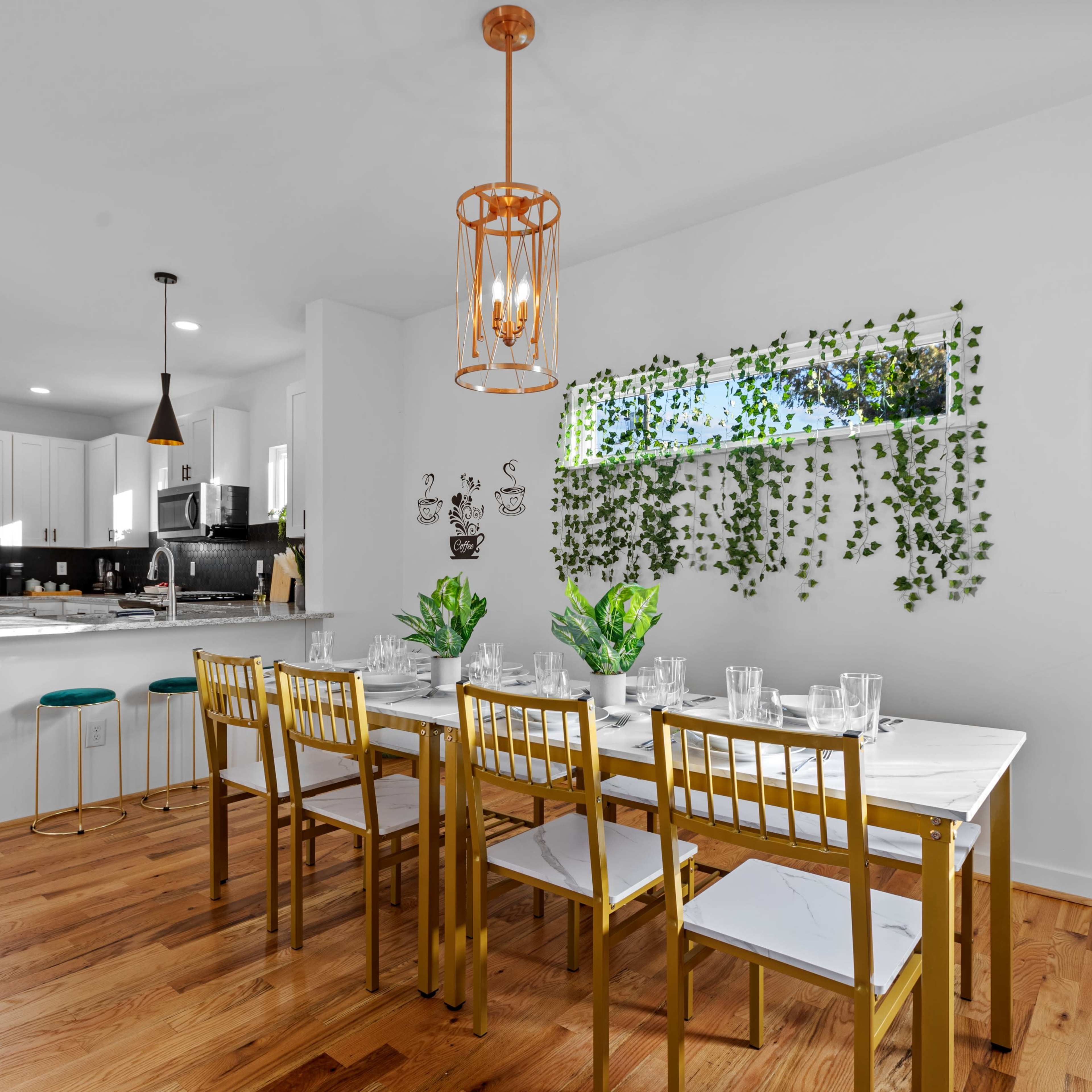 A modern dining area features a long table with gold chairs, surrounded by greenery and illuminated by pendant lights.