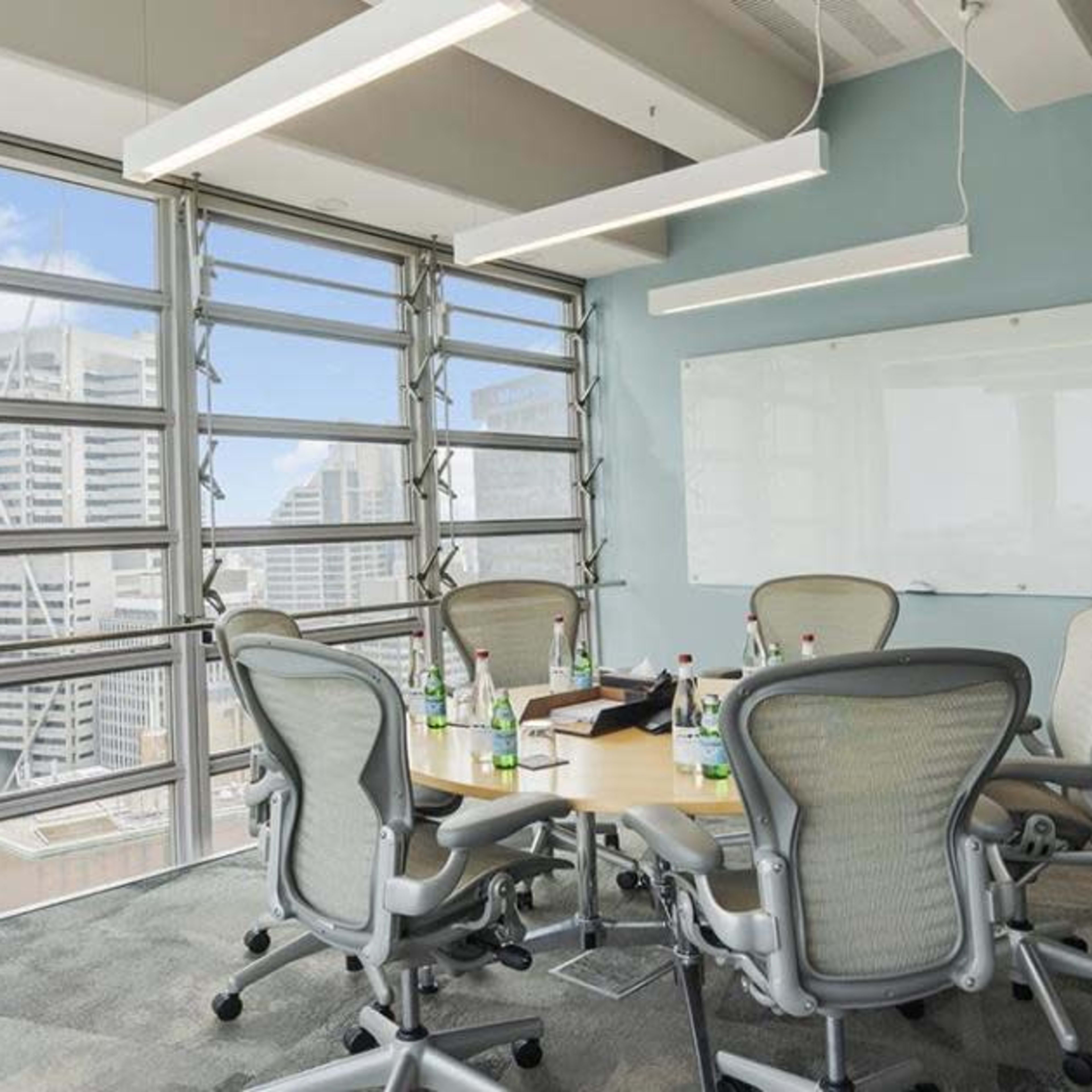 A conference room with a round table surrounded by six chairs, large windows offering a view of a city skyline, and a whiteboard on the wall.