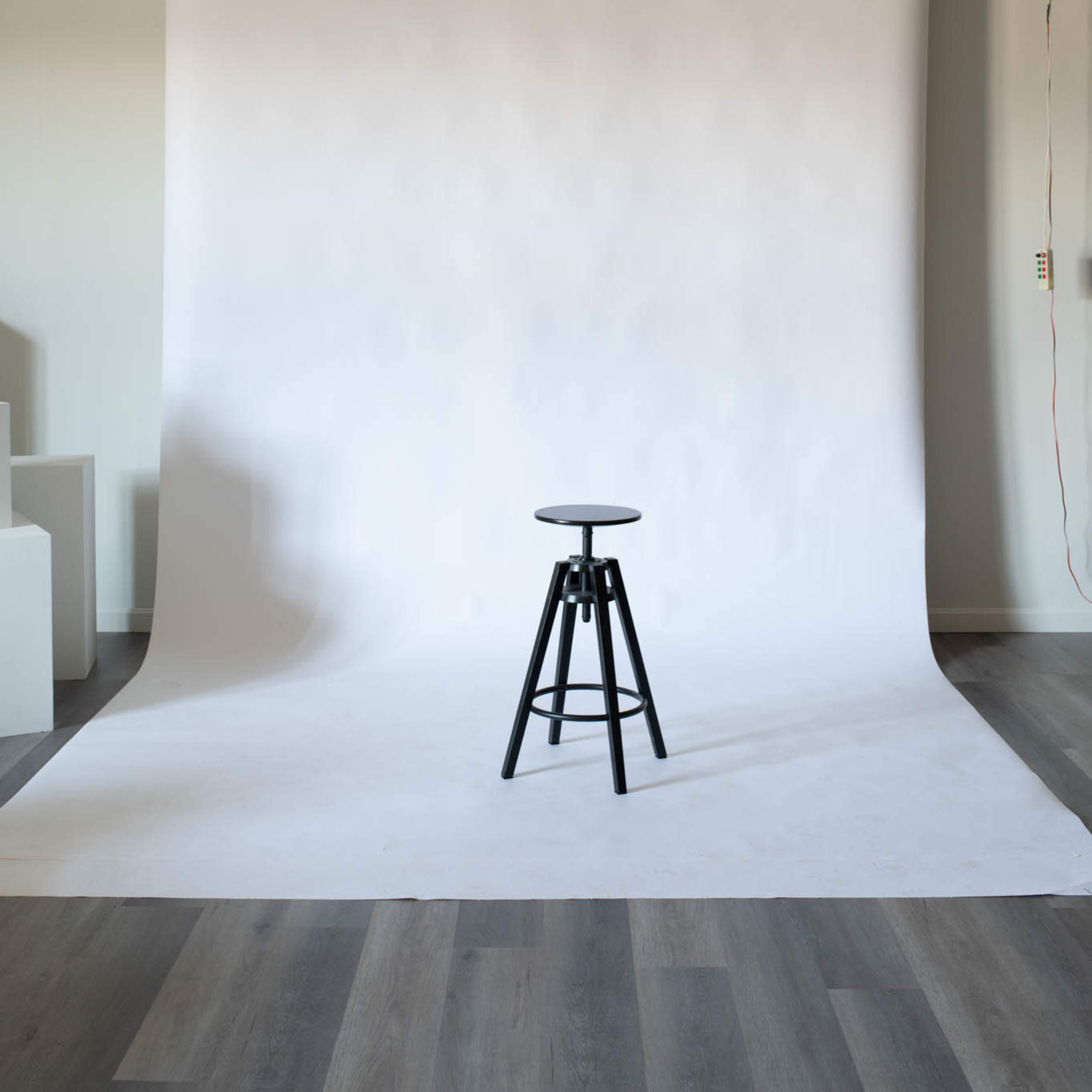 A black stool is positioned in front of a plain white backdrop in a studio setting.