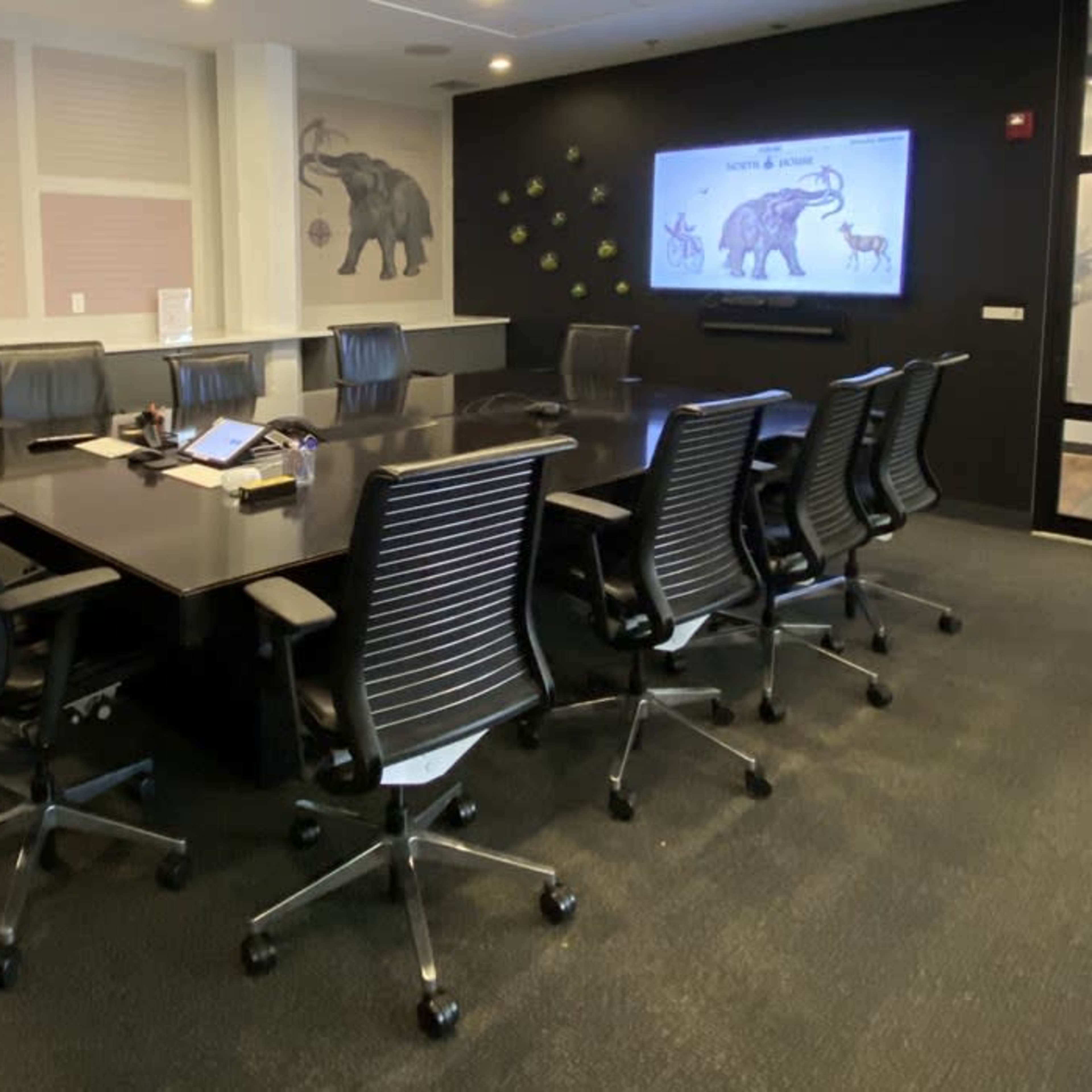 A conference room features a large black table surrounded by eight swivel chairs, with a screen displaying an elephant and walls adorned with art.