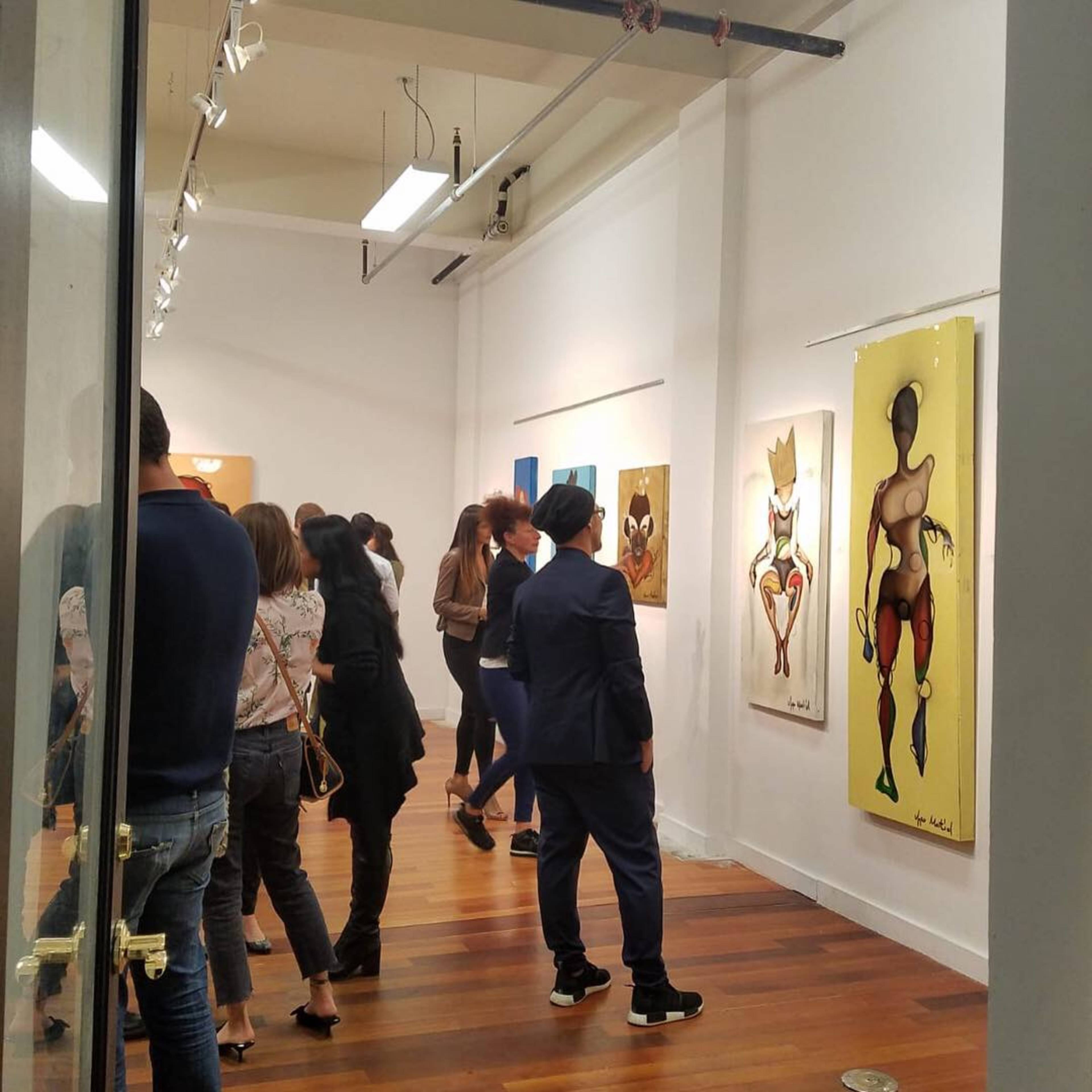 A group of people stands in an art gallery, observing various colorful paintings displayed on the walls.