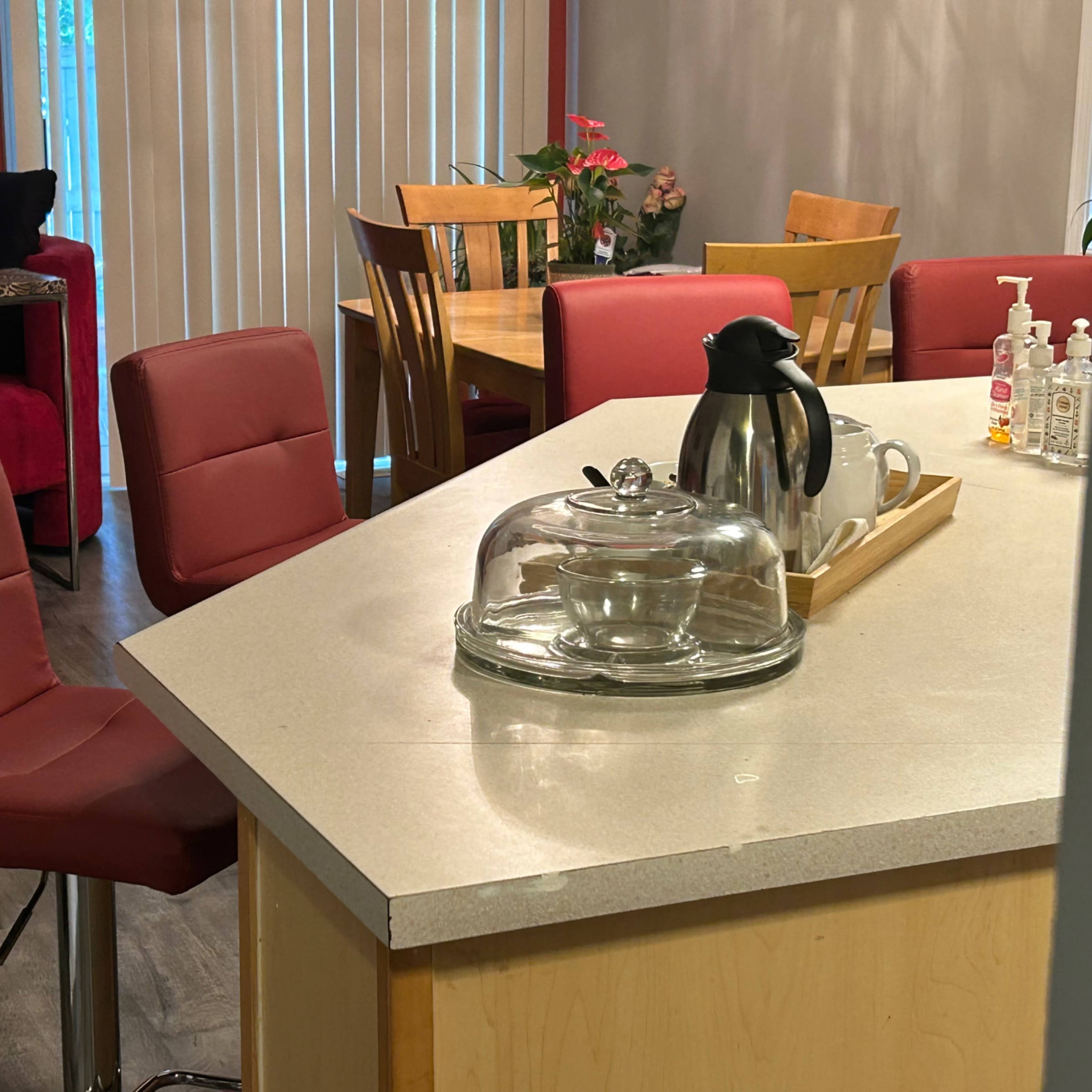 A kitchen area featuring a countertop with a glass serving dome, a coffee pot, and several red chairs arranged around the space.