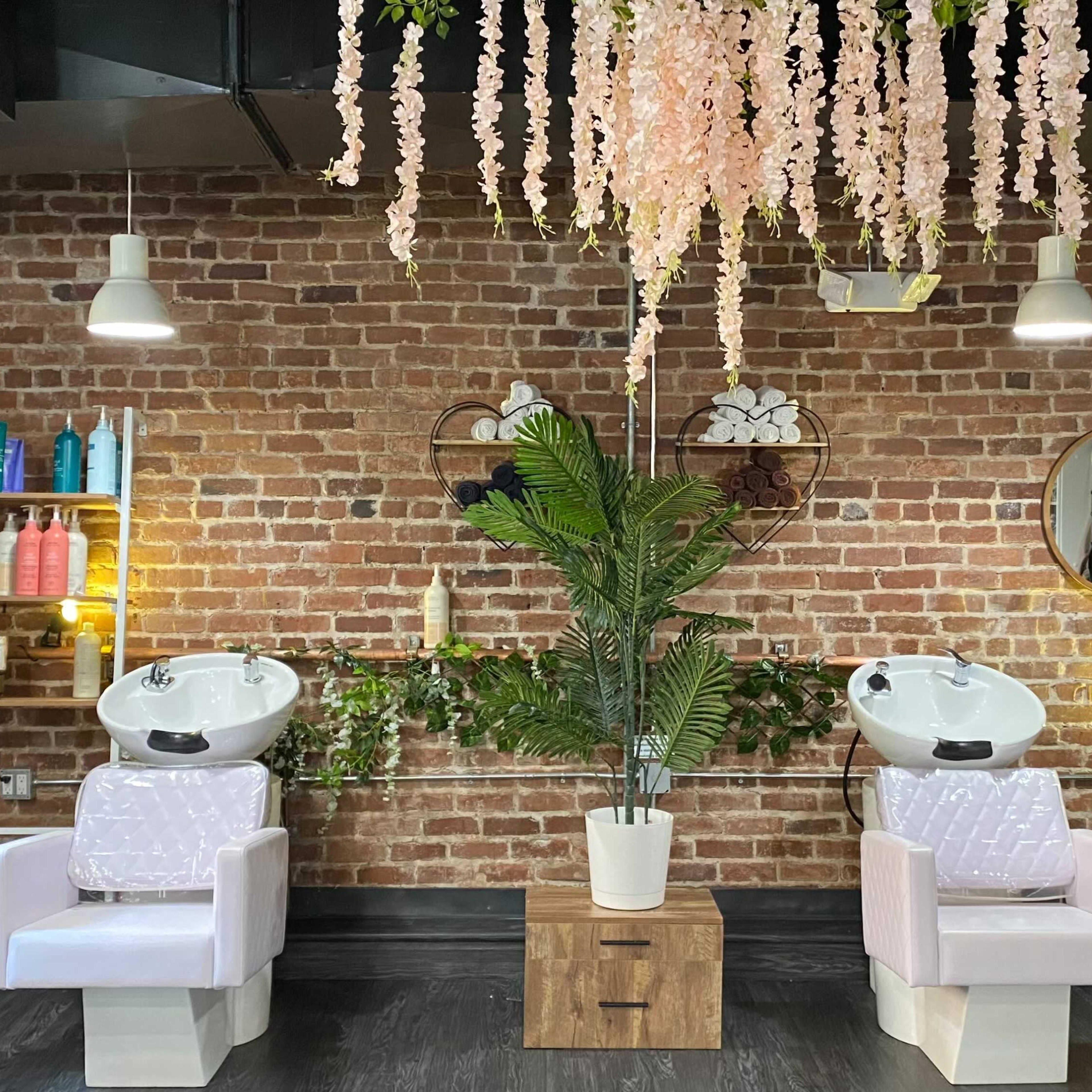 A salon interior featuring two hair washing stations with backlit mirrors, surrounded by a brick wall and adorned with hanging flowers and plants.