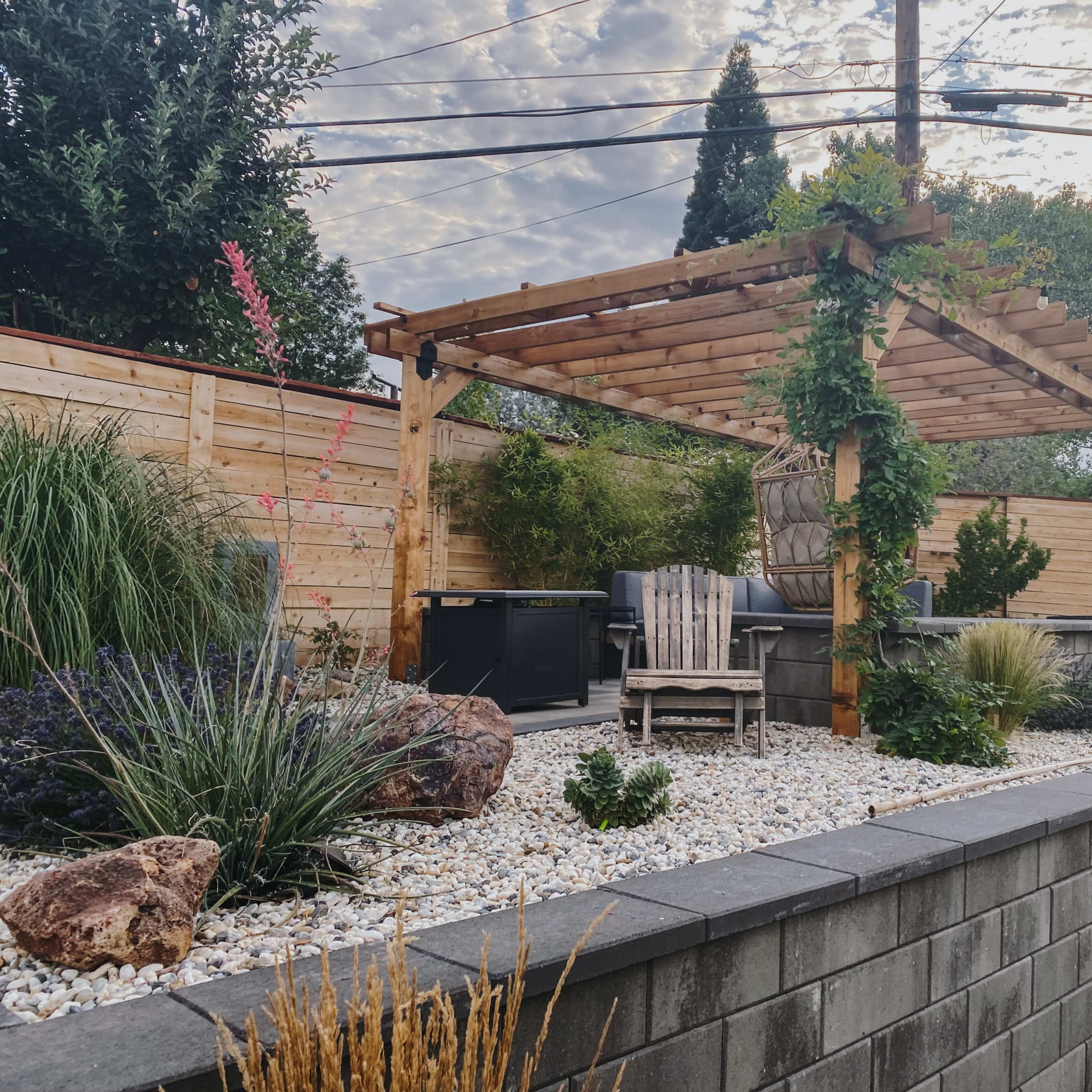 A wooden pergola with seating overlooks a landscaped garden featuring gravel, plants, and a BBQ area.