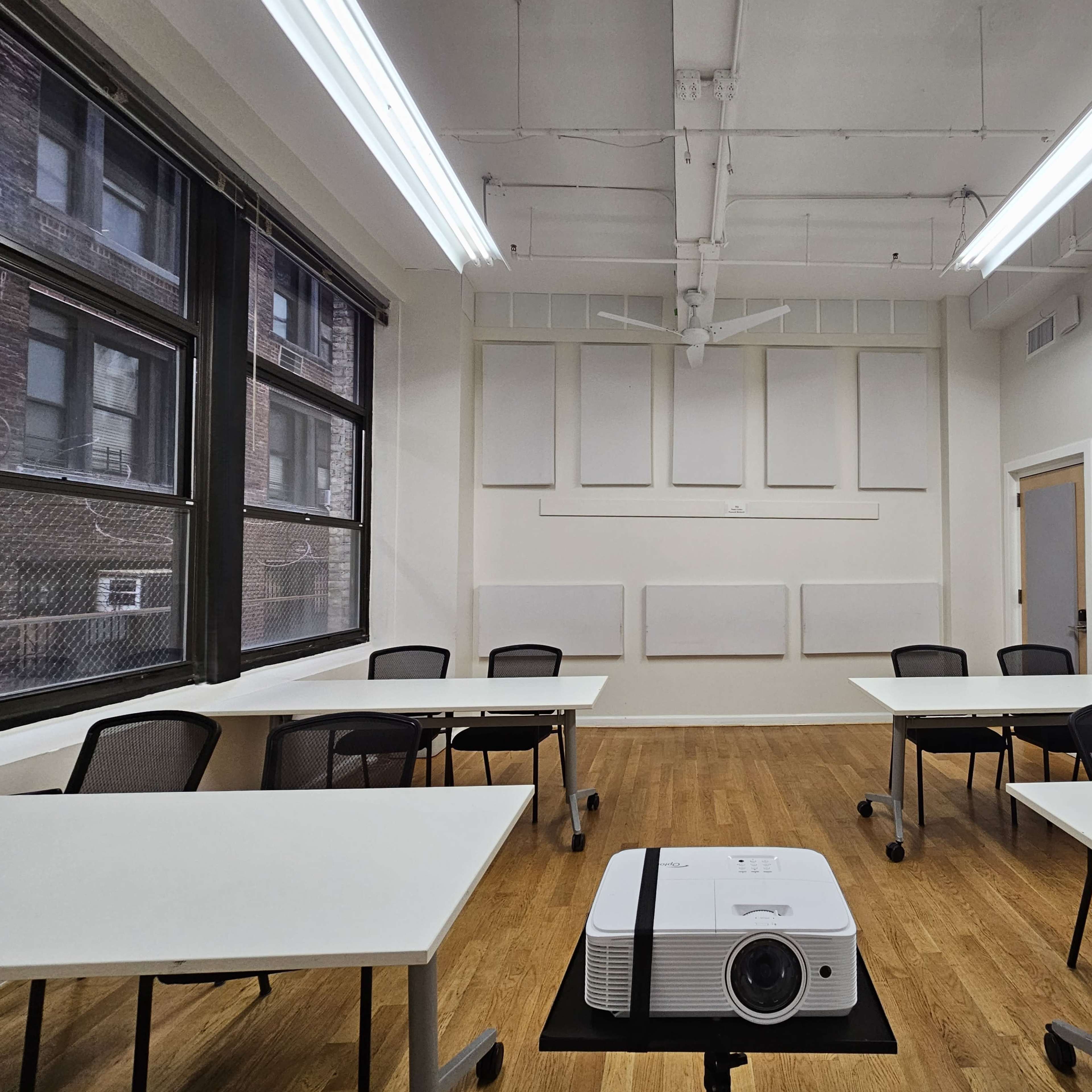 A classroom is arranged with several white tables and chairs, featuring a ceiling-mounted projector at the center.