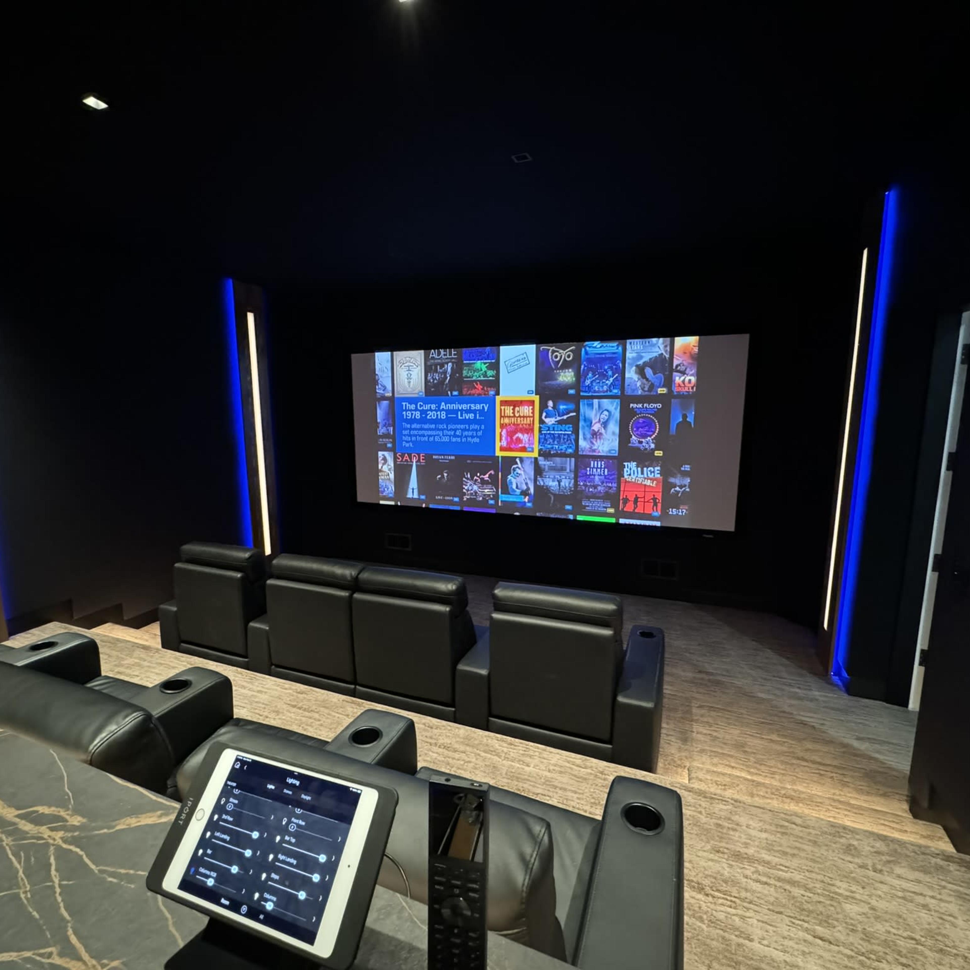 A dimly lit home theater with a large screen displaying a media library, and several black reclining seats arranged in front of it.