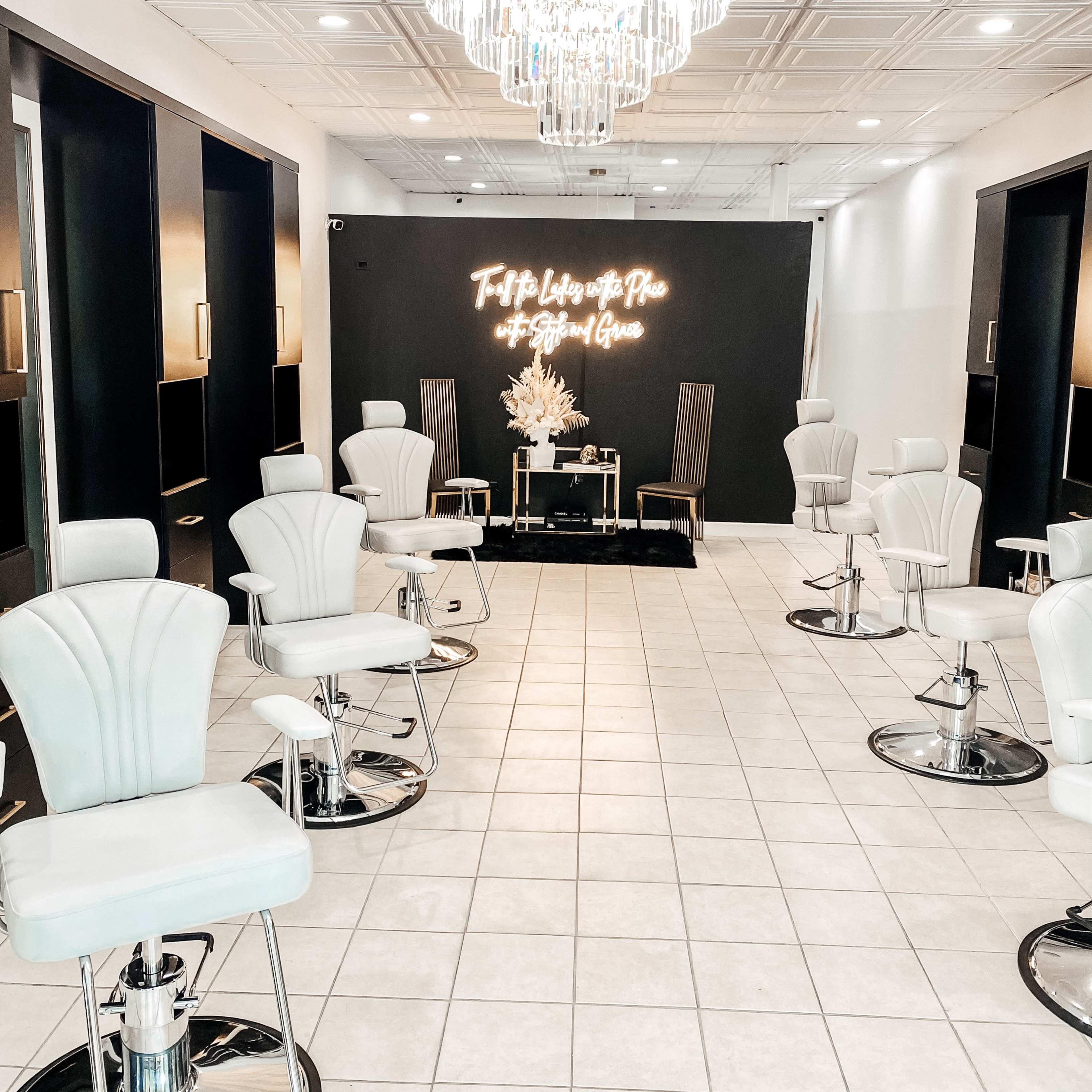 A modern salon interior featuring several white styling chairs arranged in front of a black wall with neon lighting and a chandelier above.