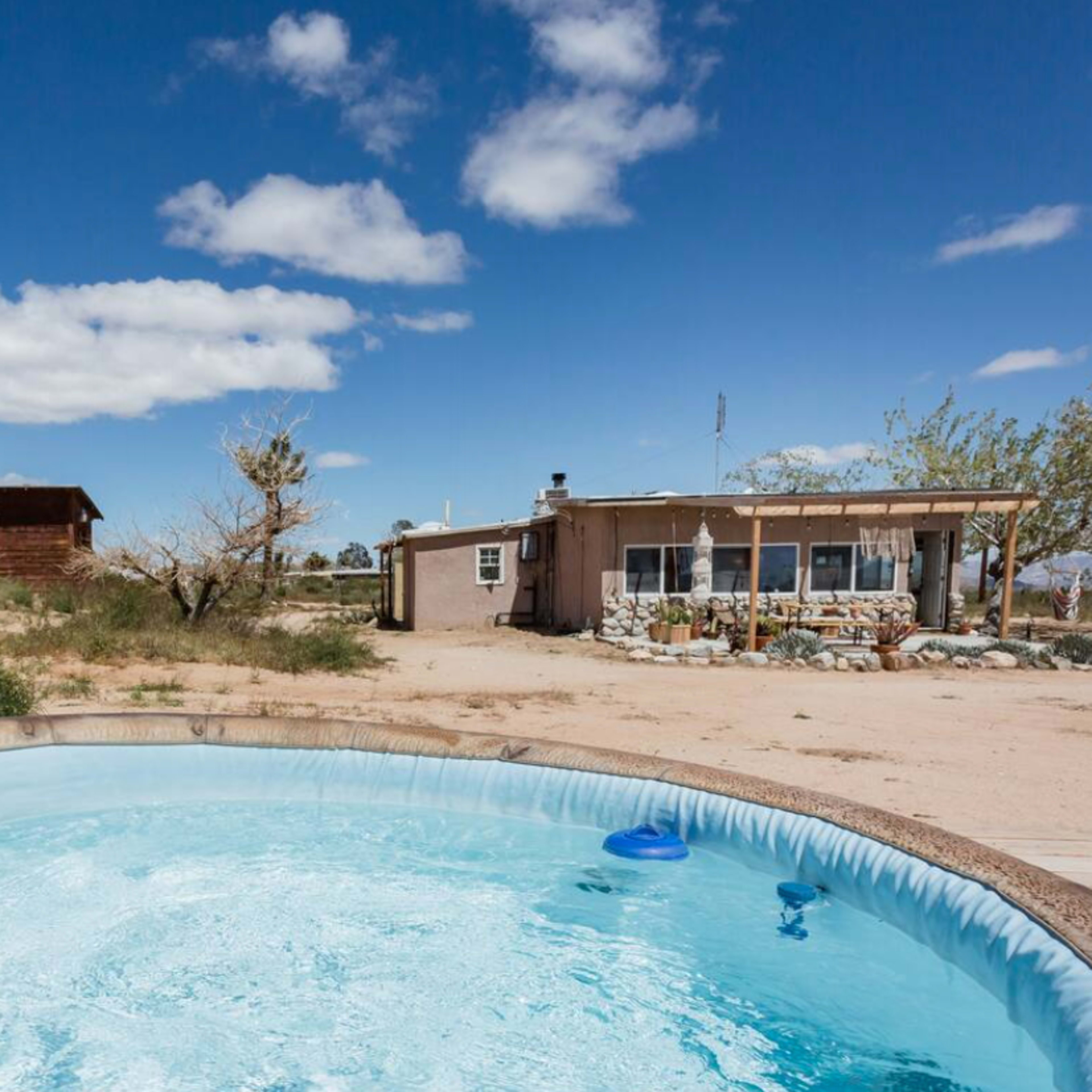 A swimming pool is visible in the foreground, with a house and desert landscape in the background under a blue sky with scattered clouds.