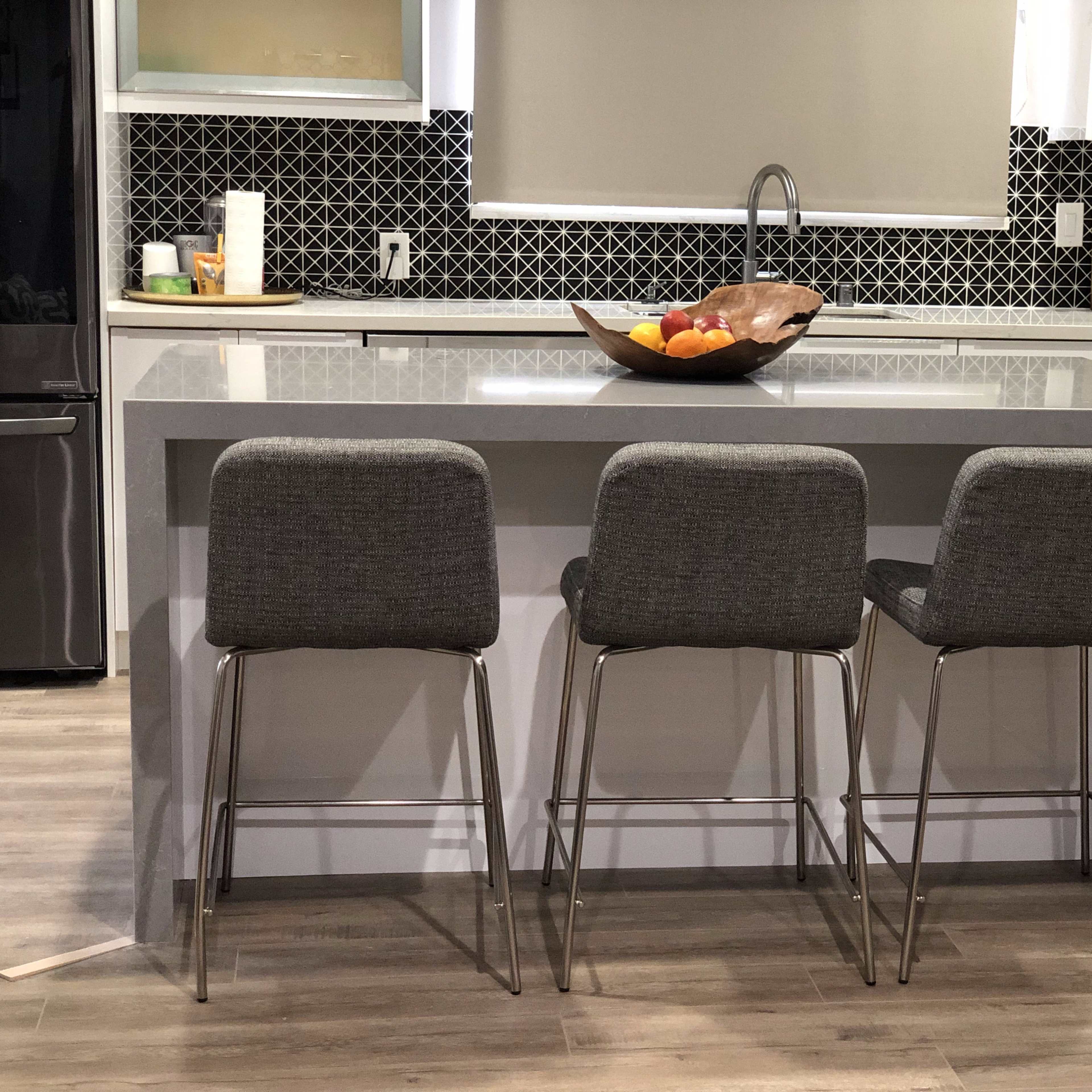 A modern kitchen with a gray island topped with a bowl of fruit and three high-backed stools arranged around it.