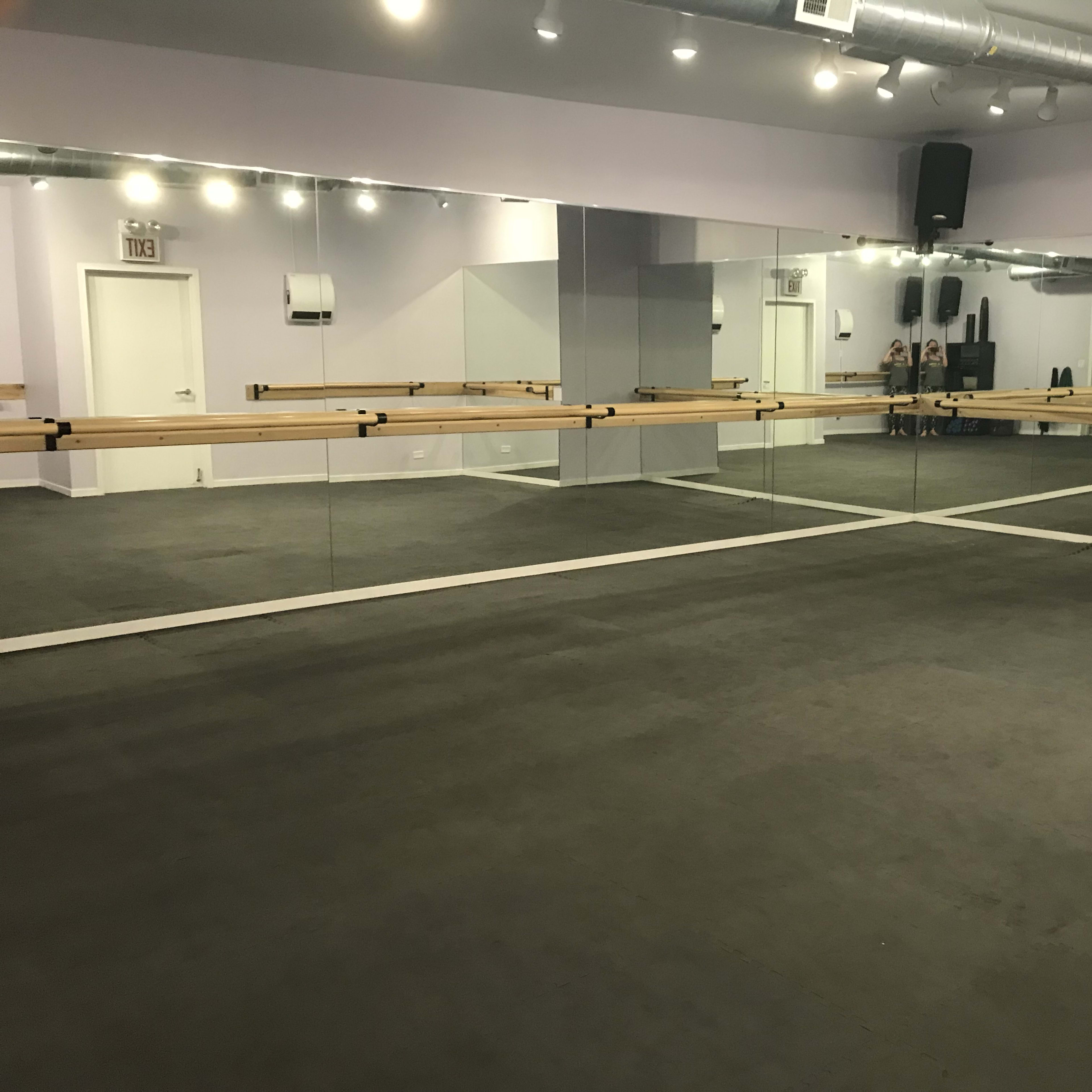 A dance studio with mirrored walls and a wooden ballet bar along one side, featuring a dark floor and bright overhead lights.