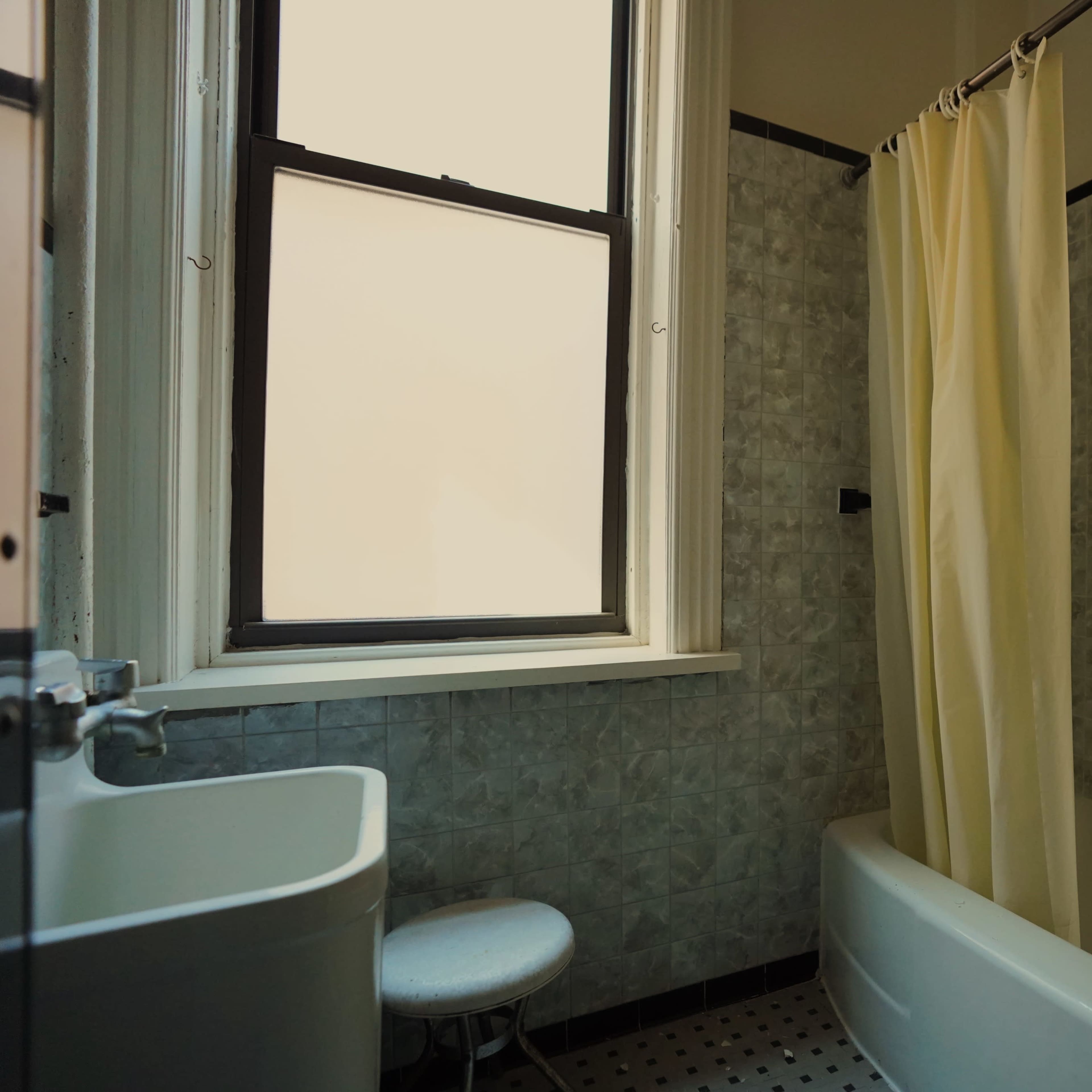A bathroom with a bathtub, a shower curtain, a small stool, and a window allowing natural light.