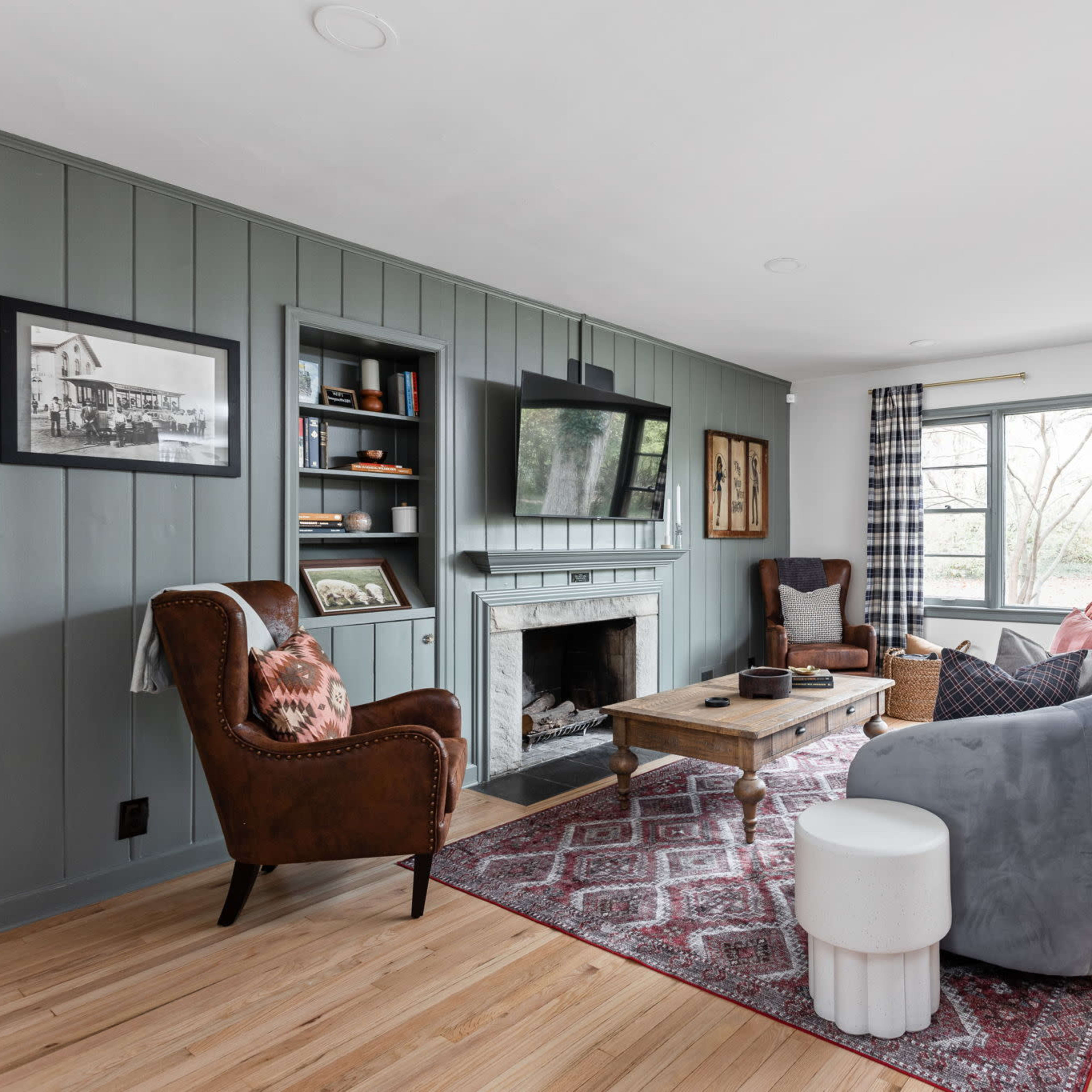 The living room features a gray sofa, two armchairs, a wooden coffee table, and a fireplace, with large windows providing natural light and a view of the outdoors.