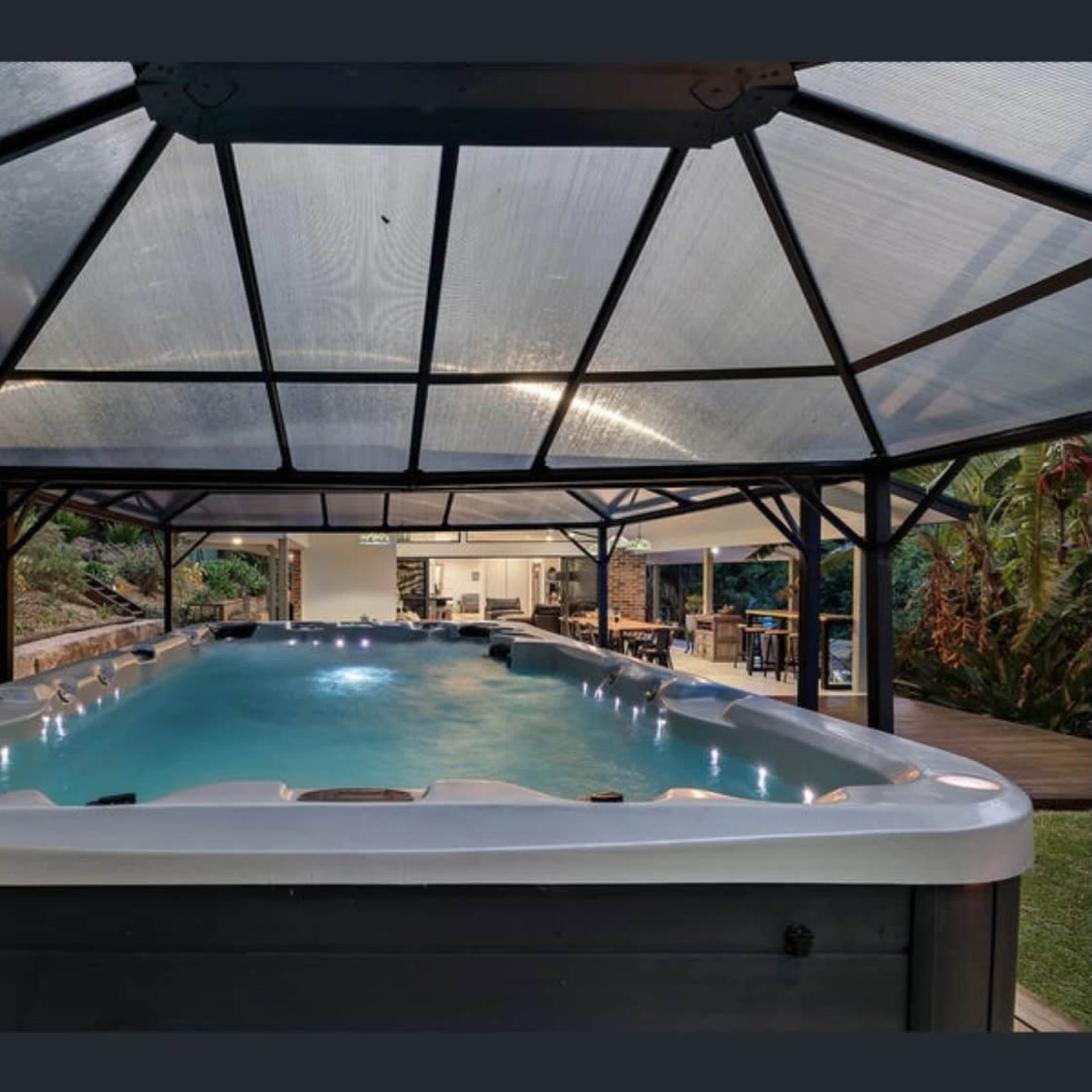A spacious indoor swimming pool housed under a glass-roofed structure, surrounded by greenery and leading to an outdoor seating area.