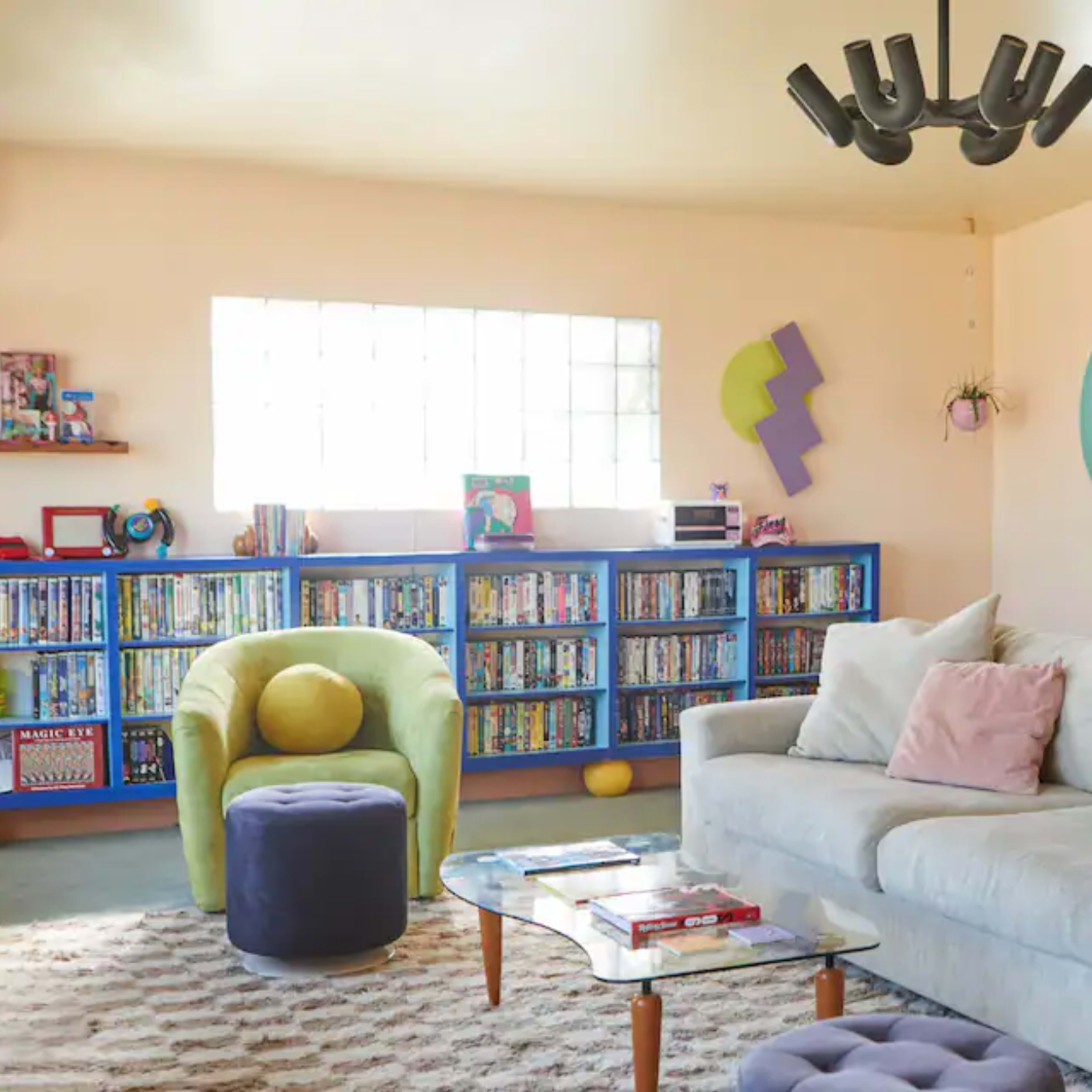 A bright living room features a light-colored sofa, colorful bookshelves filled with DVDs, and a round window, all anchored by a light fixture.