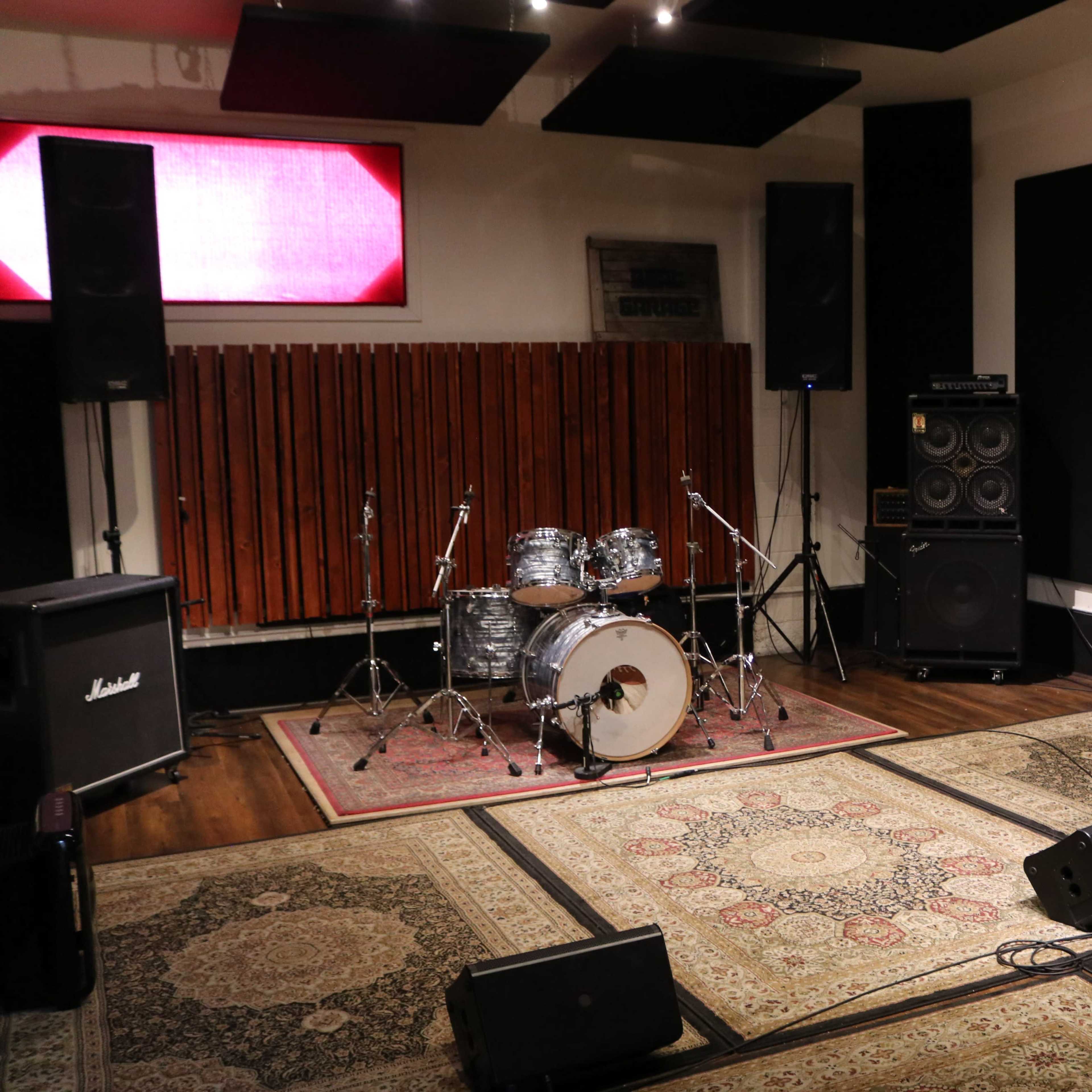 A music rehearsal room features a drum set, amplifiers, and sound equipment arranged on patterned rugs.