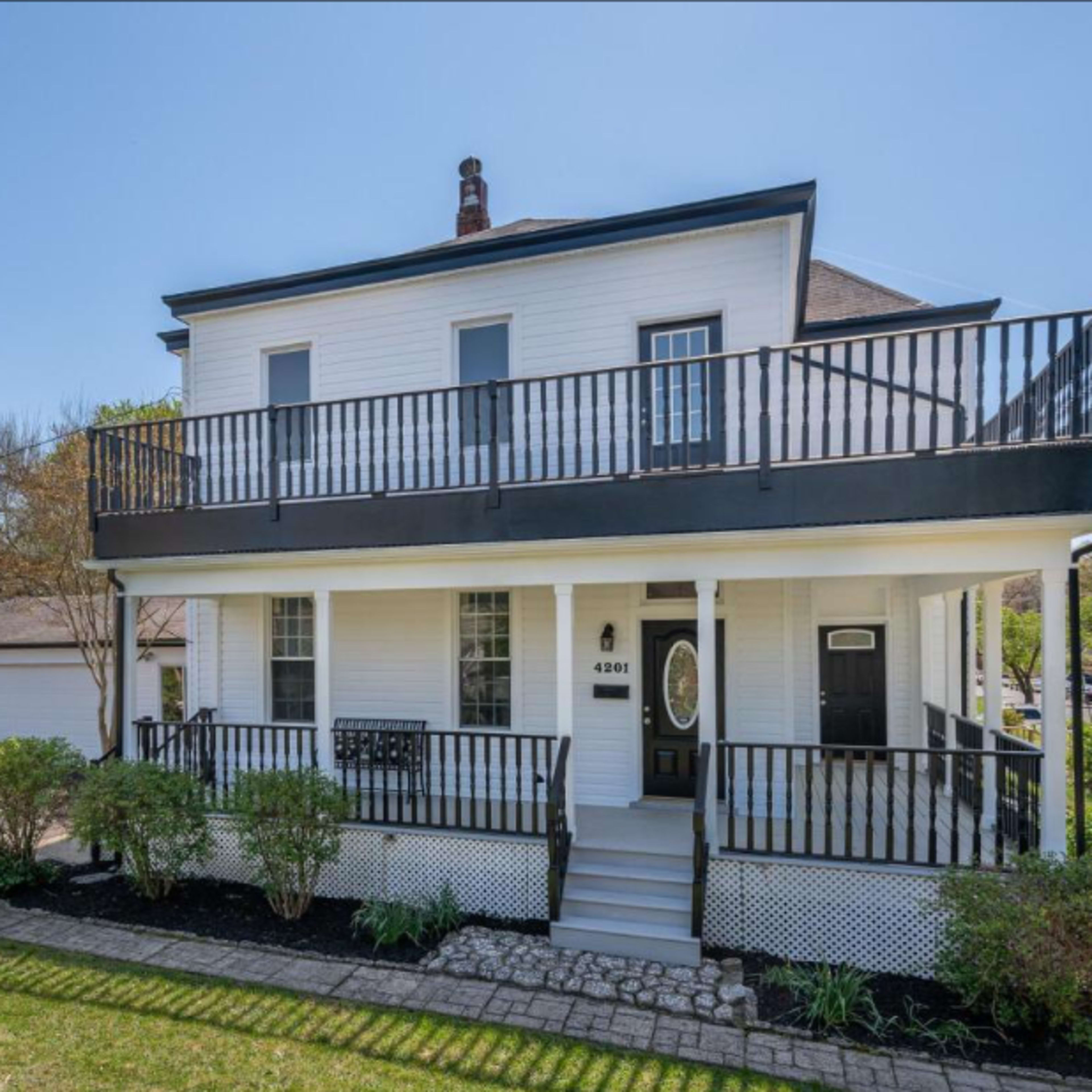 A two-story white house with a black trim and a balcony, surrounded by greenery and a driveway leading to a garage.