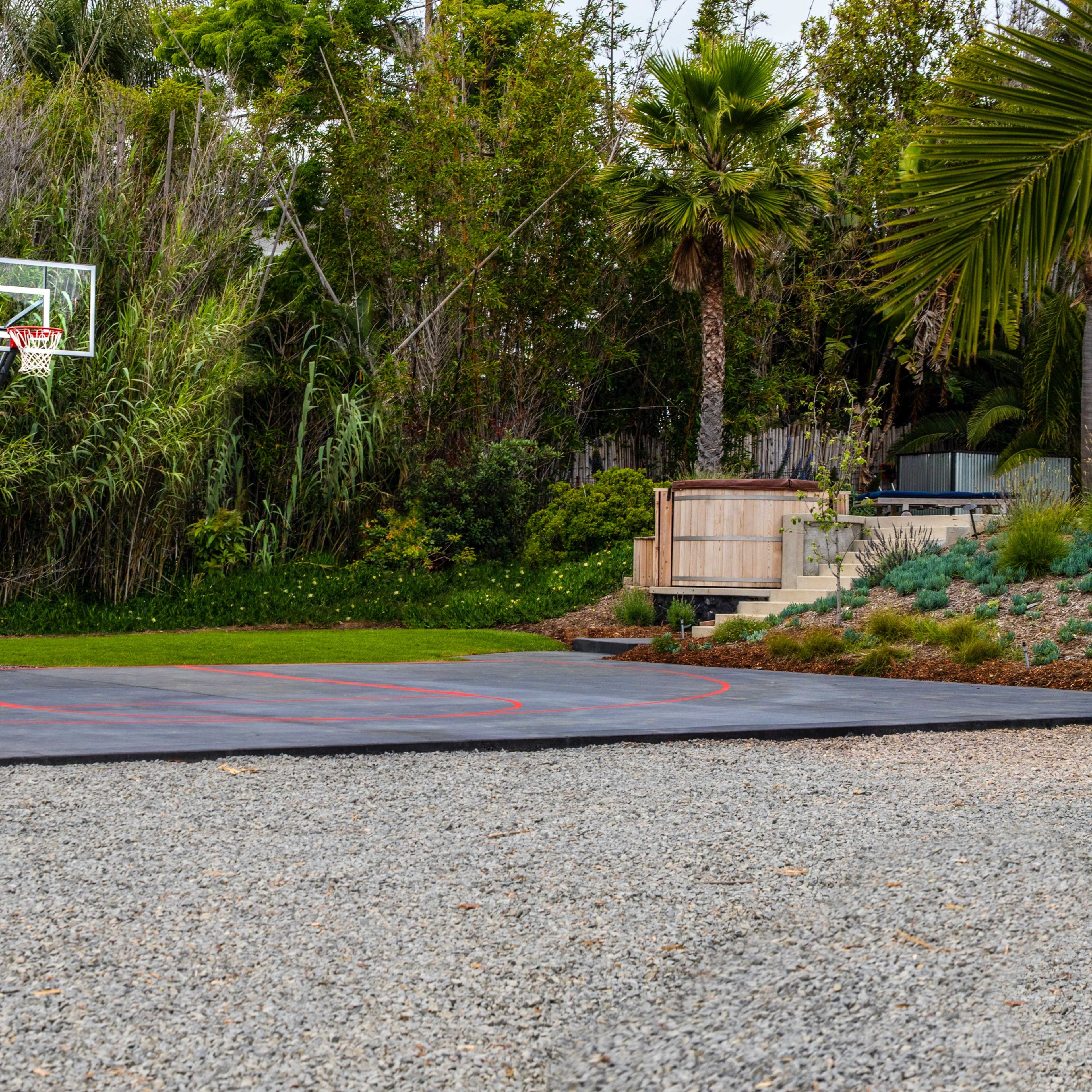 A basketball court with a hoop stands on gravel, surrounded by lush greenery and palm trees.