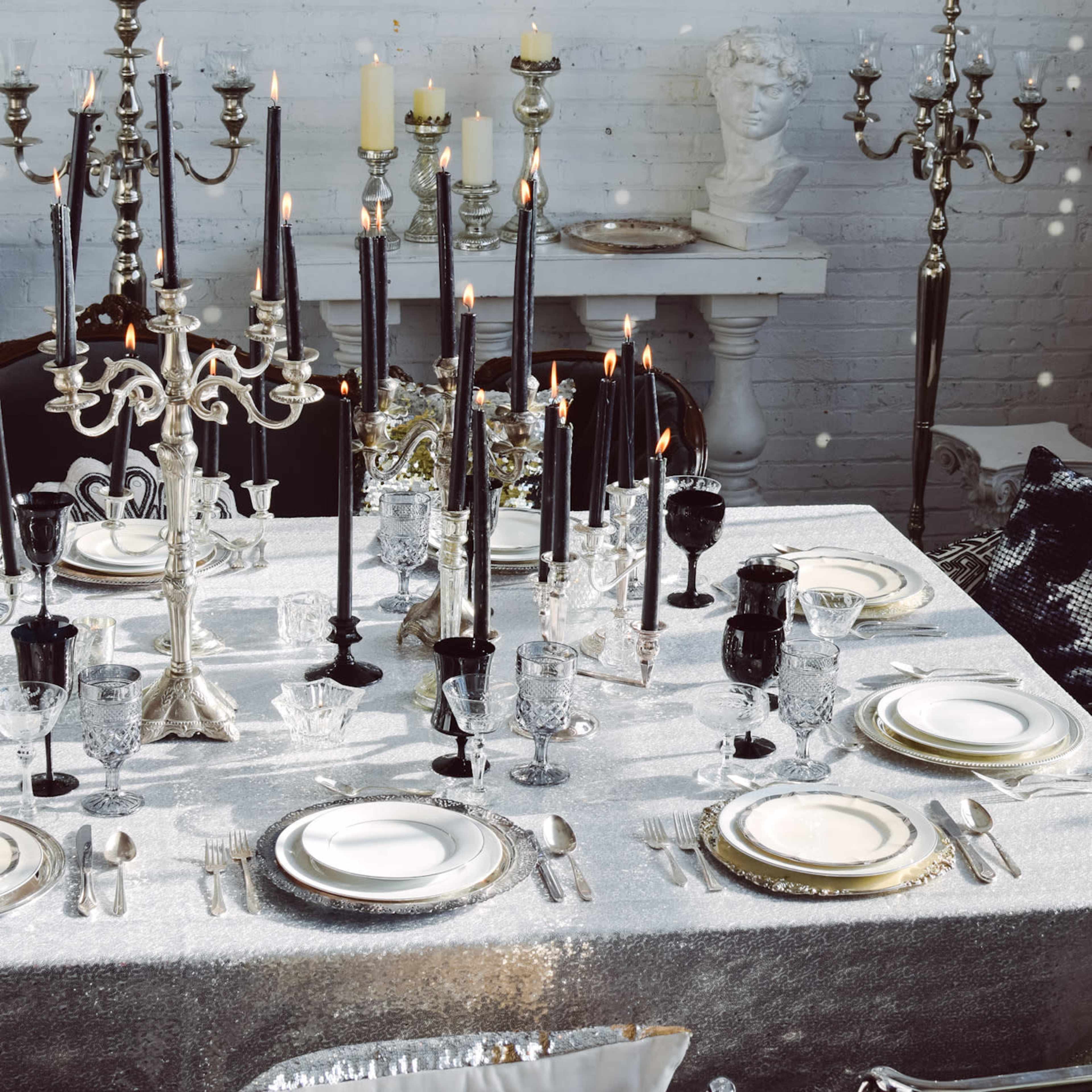 A formal dining table is elegantly set with fine china, silverware, and numerous tall candles in ornate holders, surrounded by a sleek, silver tablecloth and decorative candelabras.