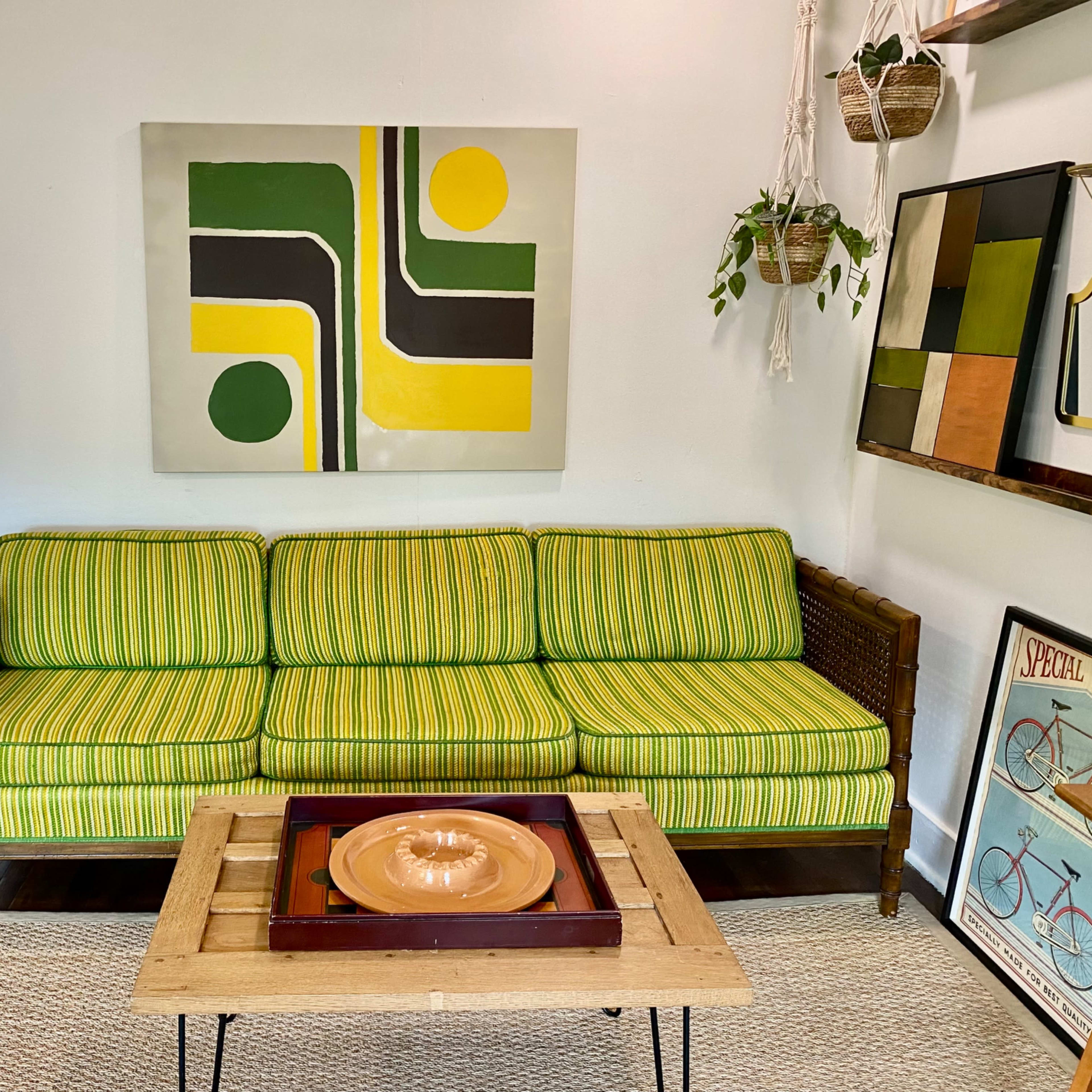 A striped green couch is positioned in a room featuring a modern abstract painting, a wooden coffee table, and potted plants hanging on the wall.