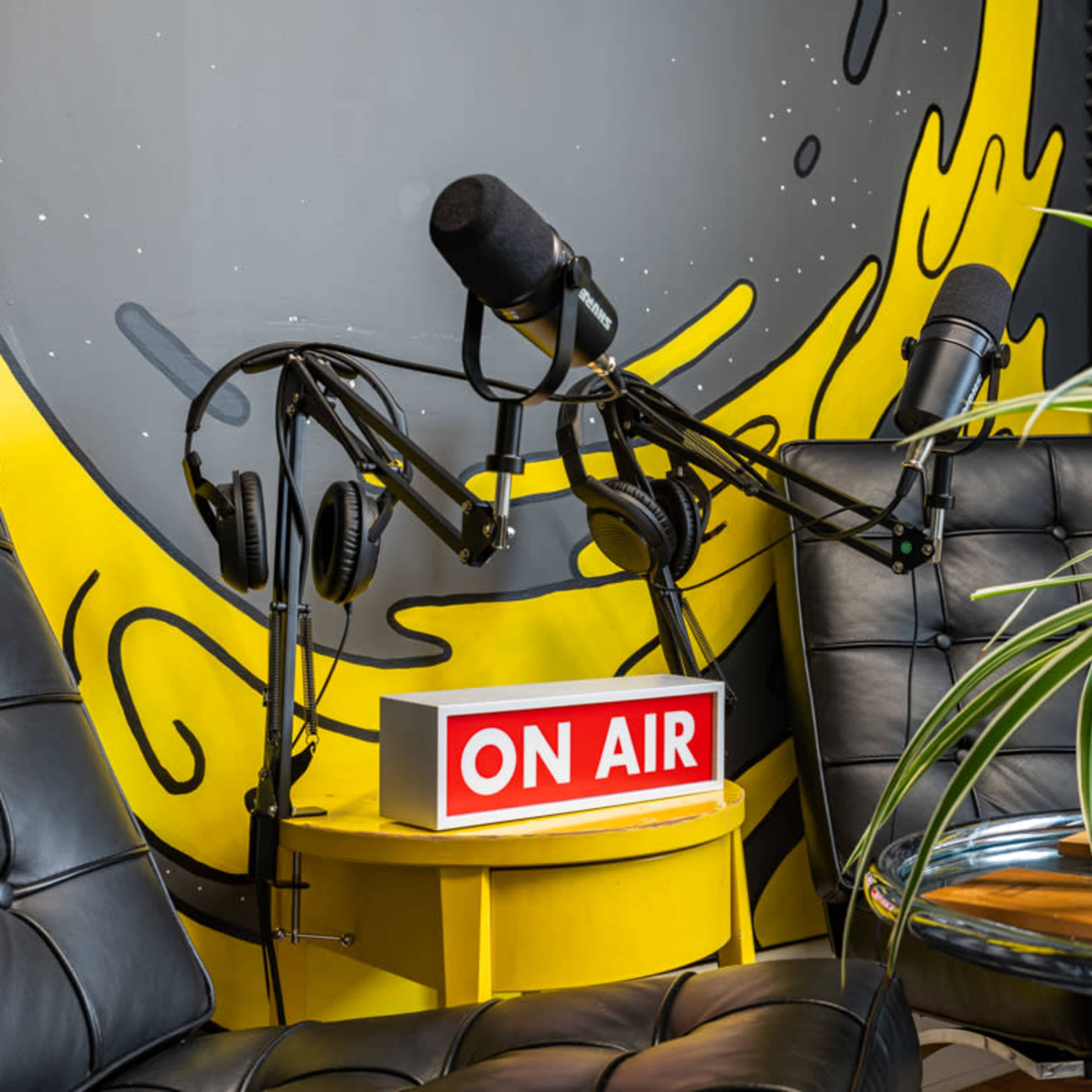 A recording studio setup with two black chairs, a "ON AIR" sign on a yellow table, and two microphones on adjustable arms beside headphones.