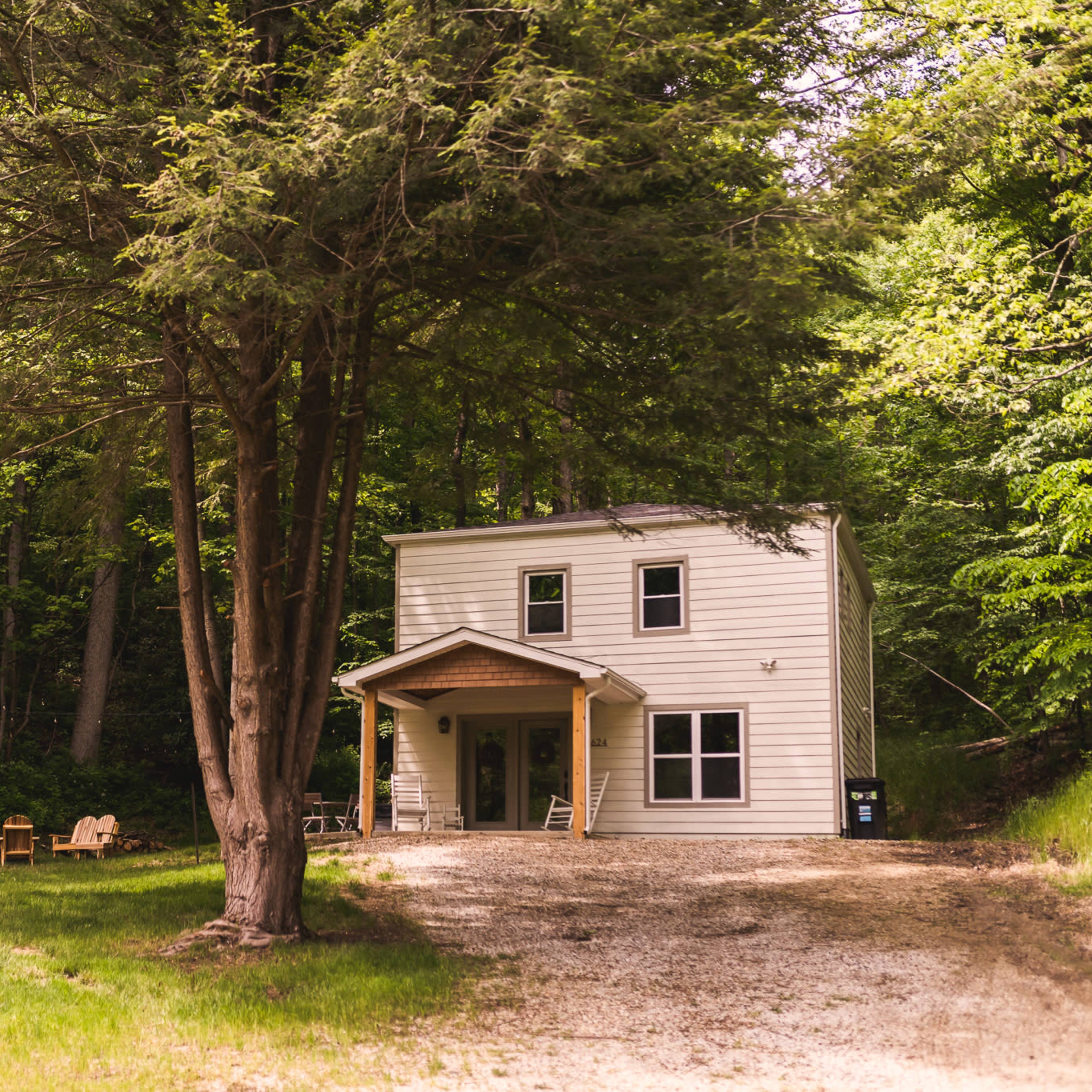 A two-story house with a porch is situated beside a gravel driveway and surrounded by trees in a forested area.