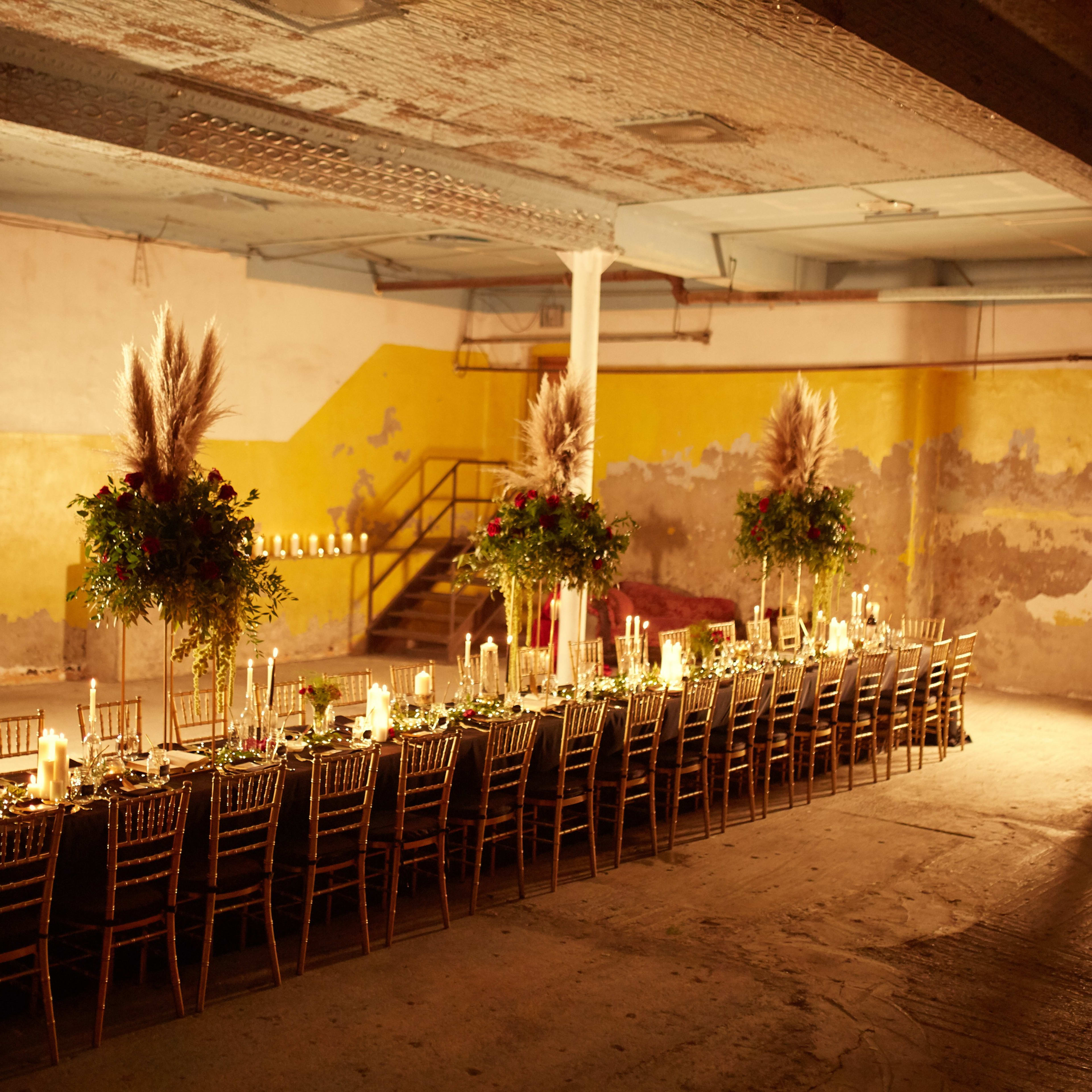 A long dining table is set in an industrial space with yellow walls and dim lighting, adorned with candles and floral arrangements.