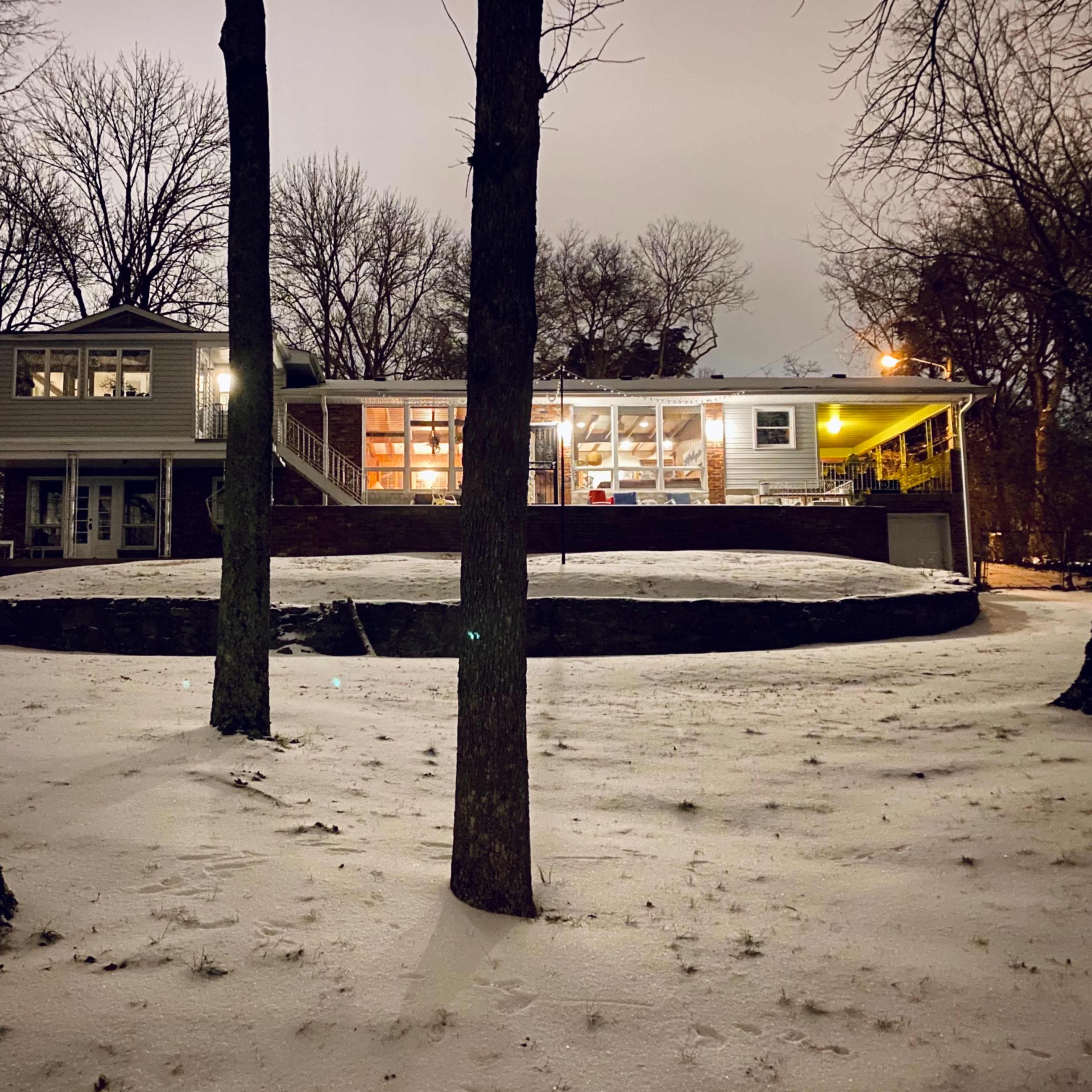 A house with large windows sits on a snow-covered lawn, surrounded by bare trees at night.