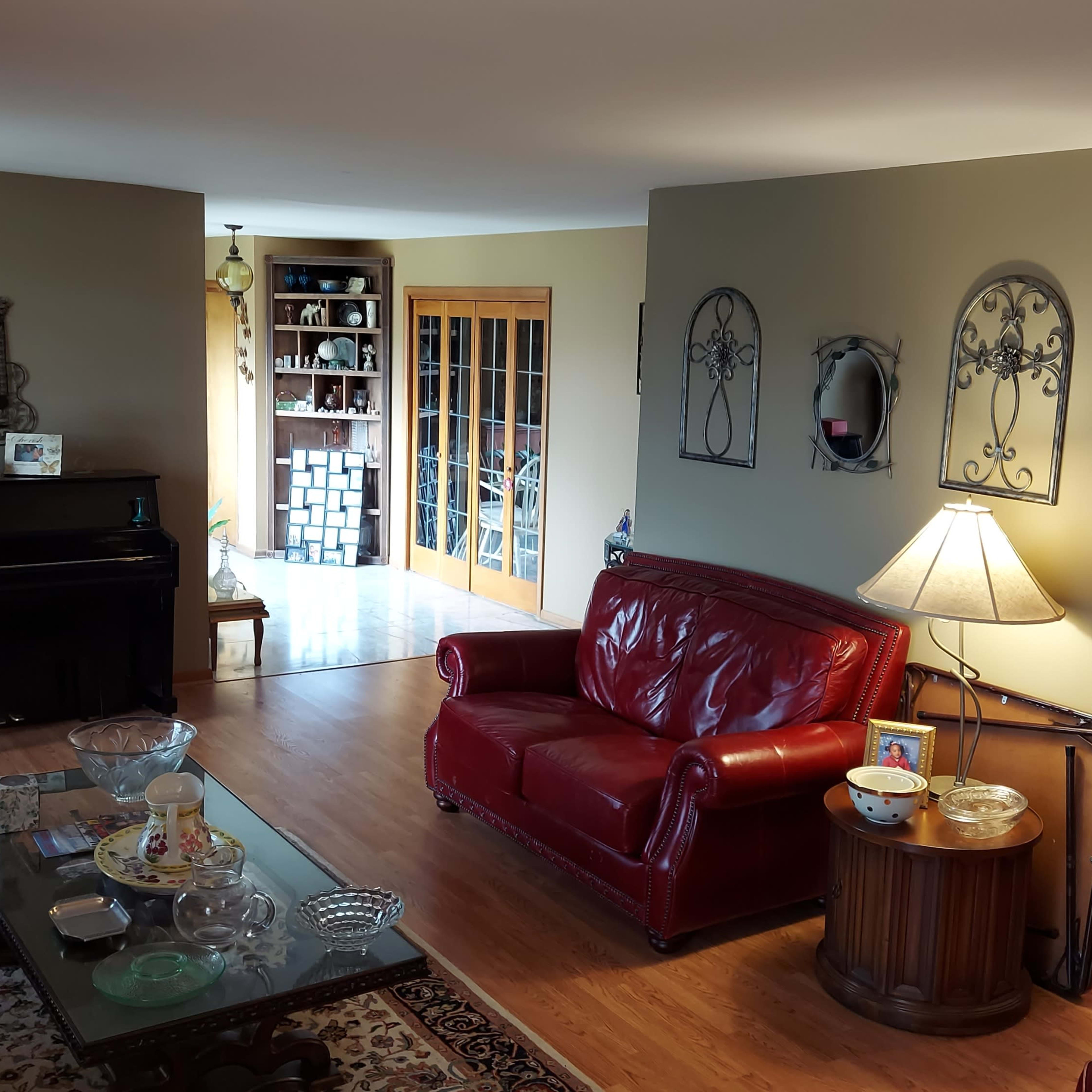A living room with a red leather sofa, a glass coffee table, a piano, and an adjoining dining area visible through double doors.