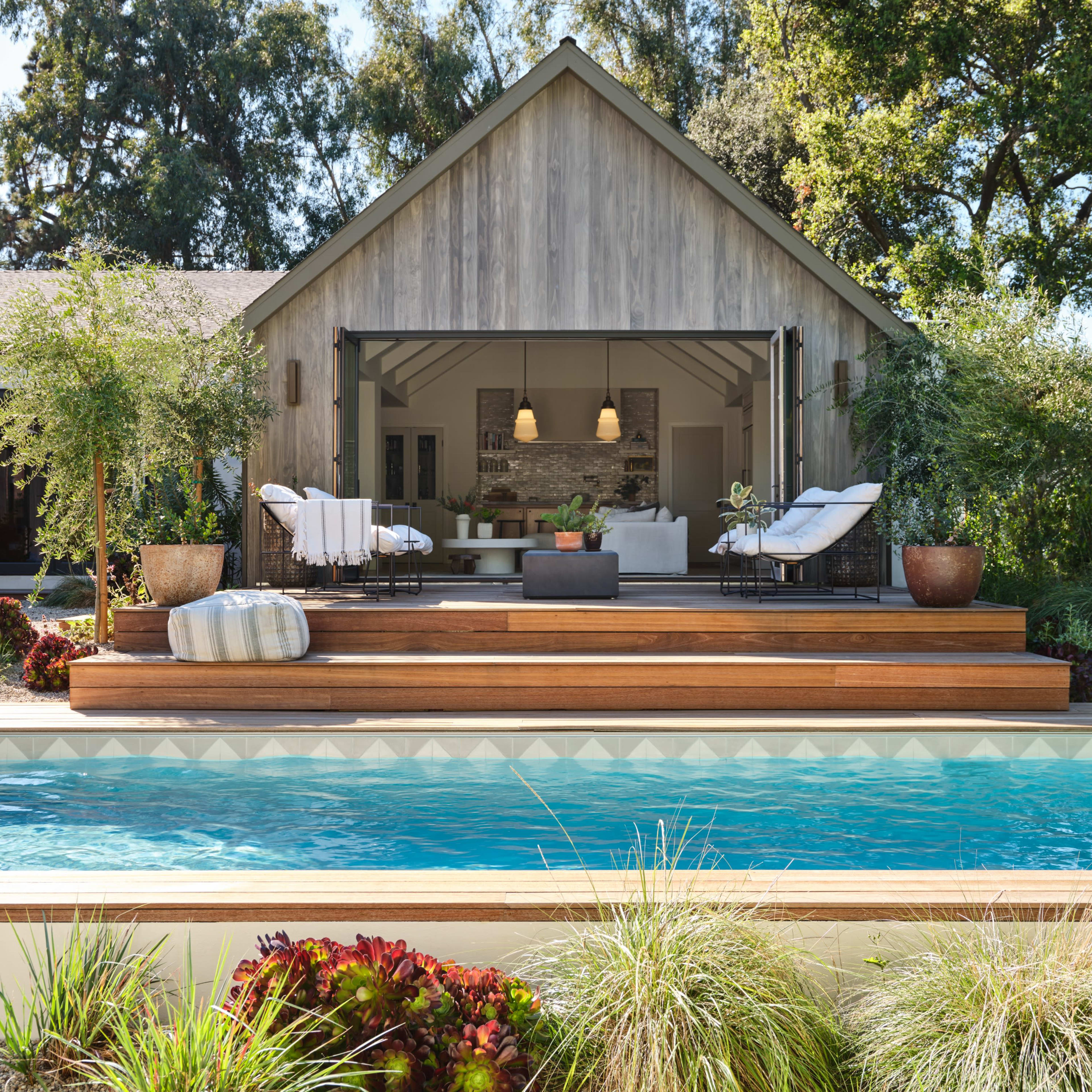 A large wooden deck with seating overlooks a swimming pool, adjacent to a modern house framed by trees.