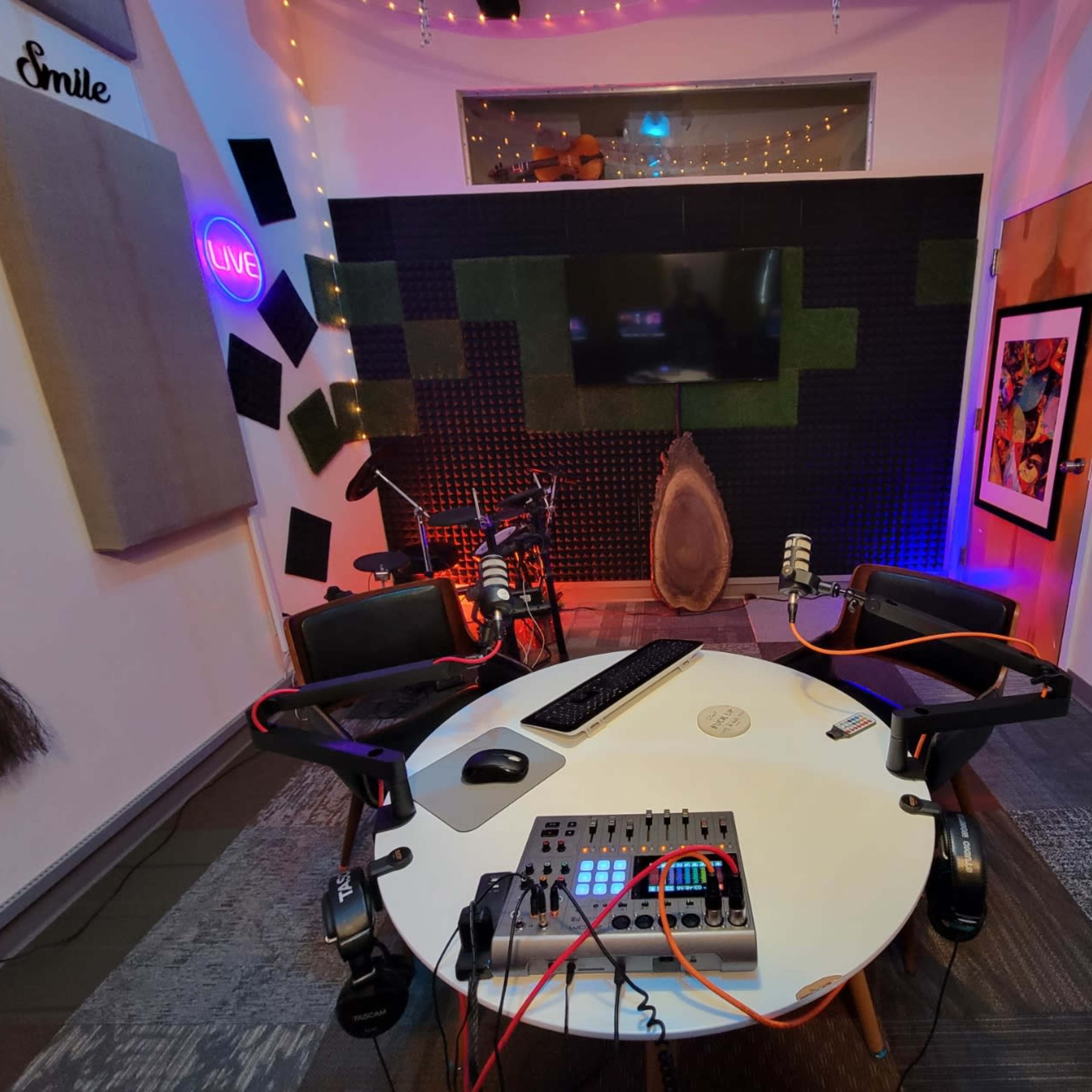 A recording studio setup featuring a circular table with microphones, a computer, and colorful lighting against a backdrop of soundproofing panels and a large screen.