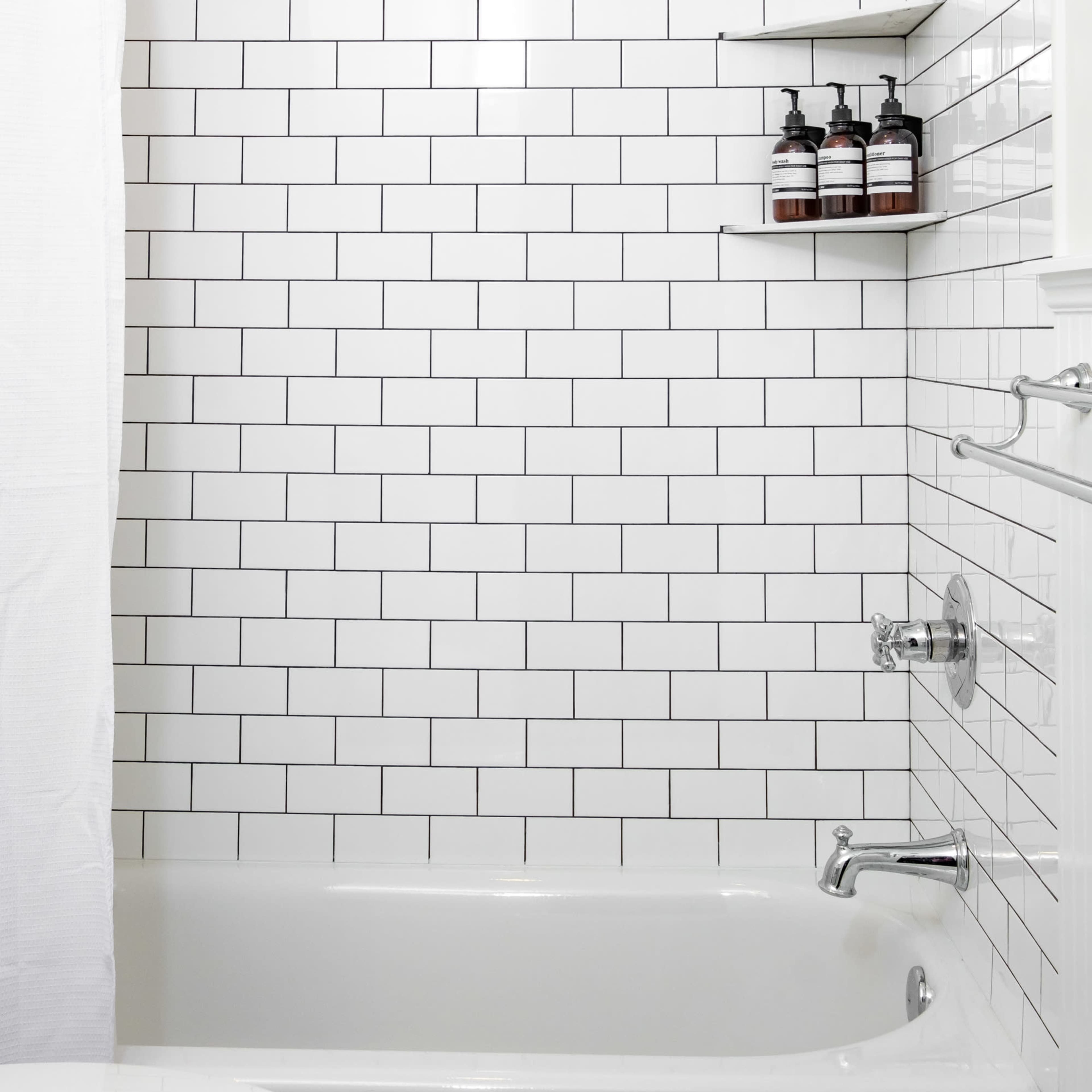 A clean, tiled shower area with a white curtain, a showerhead, and shelves holding bottles.