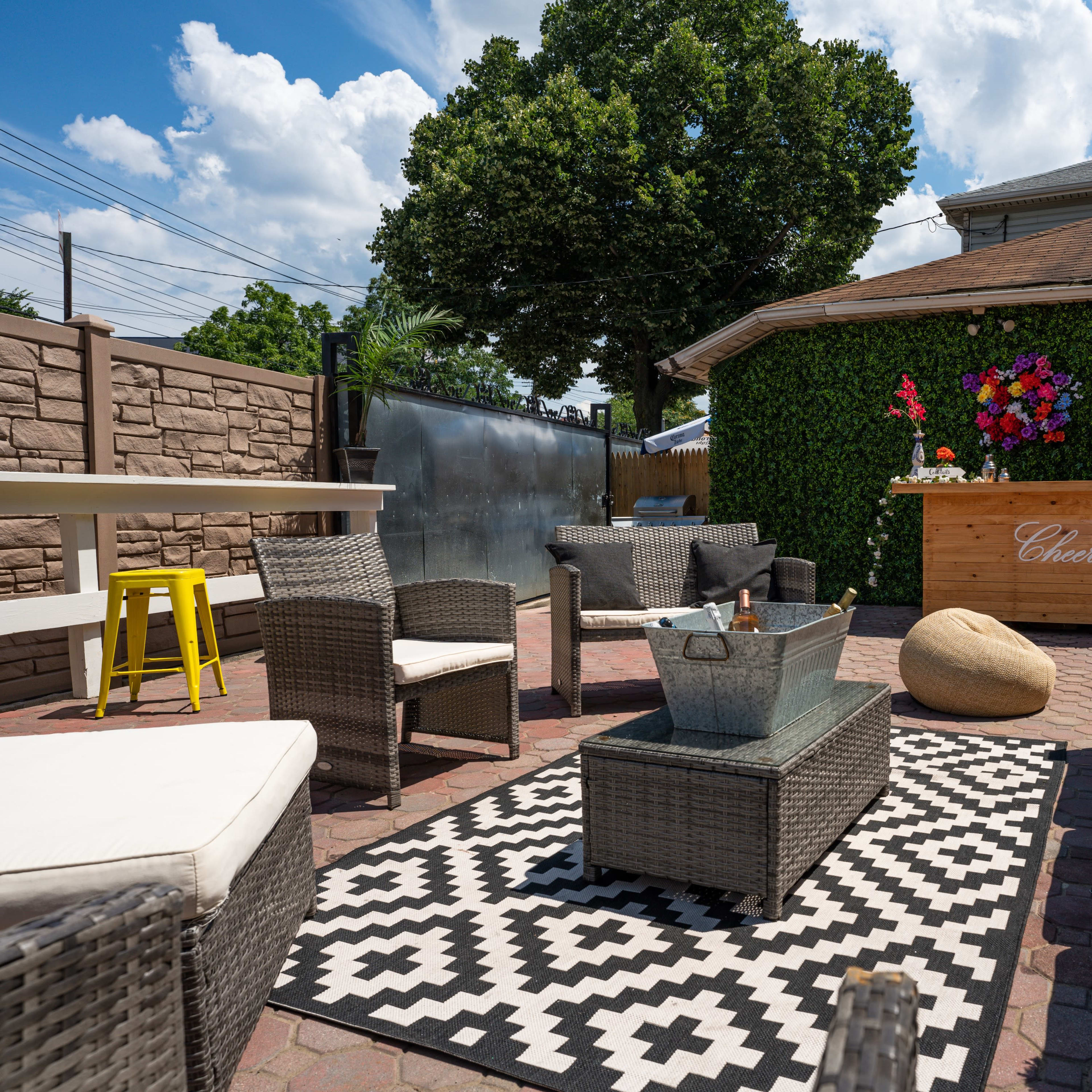 A patio area with seating arrangements, a patterned rug, a fire pit, and a bar decorated with flowers against a backdrop of green foliage.