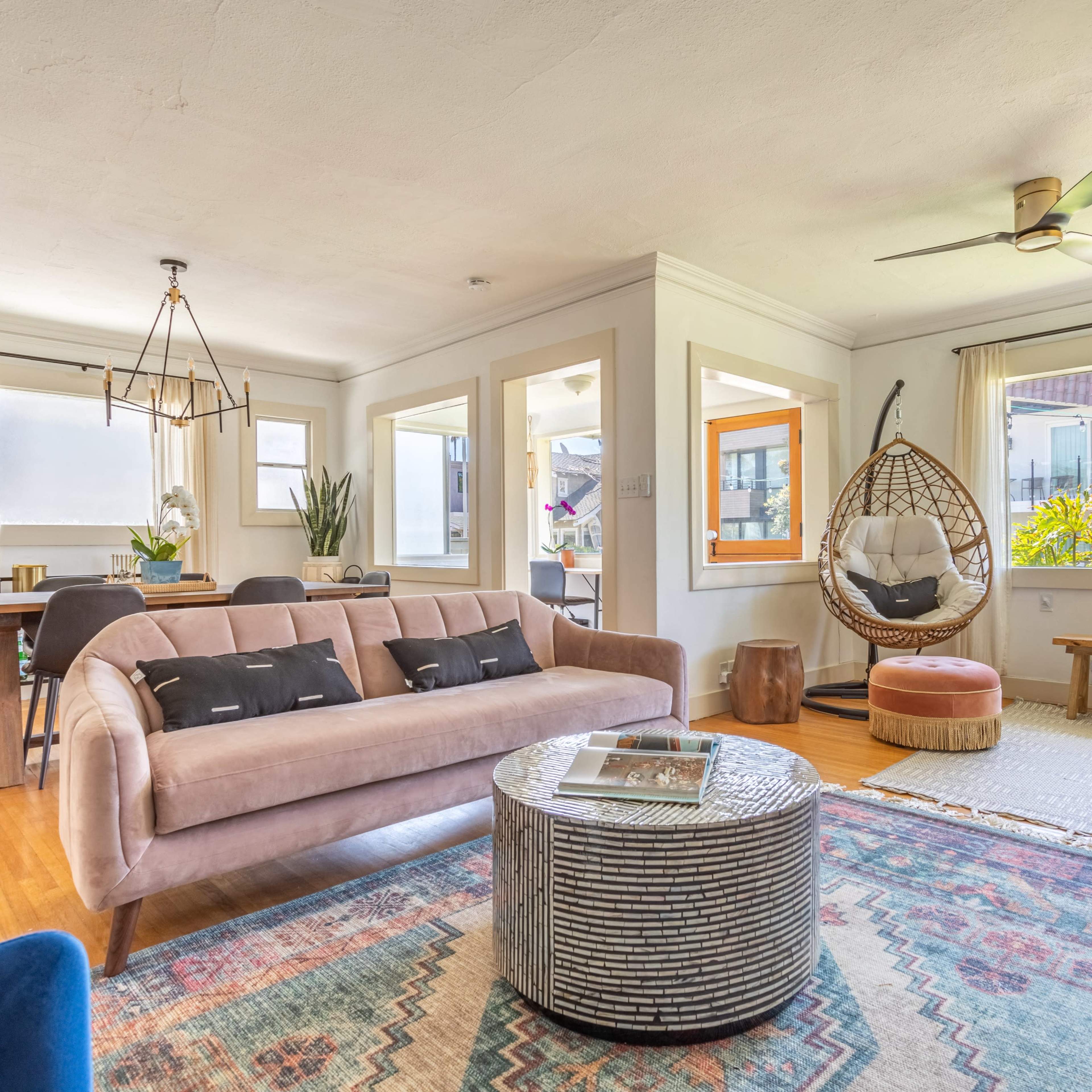 A well-furnished living room features a pink sofa, a round metallic coffee table, a hanging chair, and large windows allowing natural light.