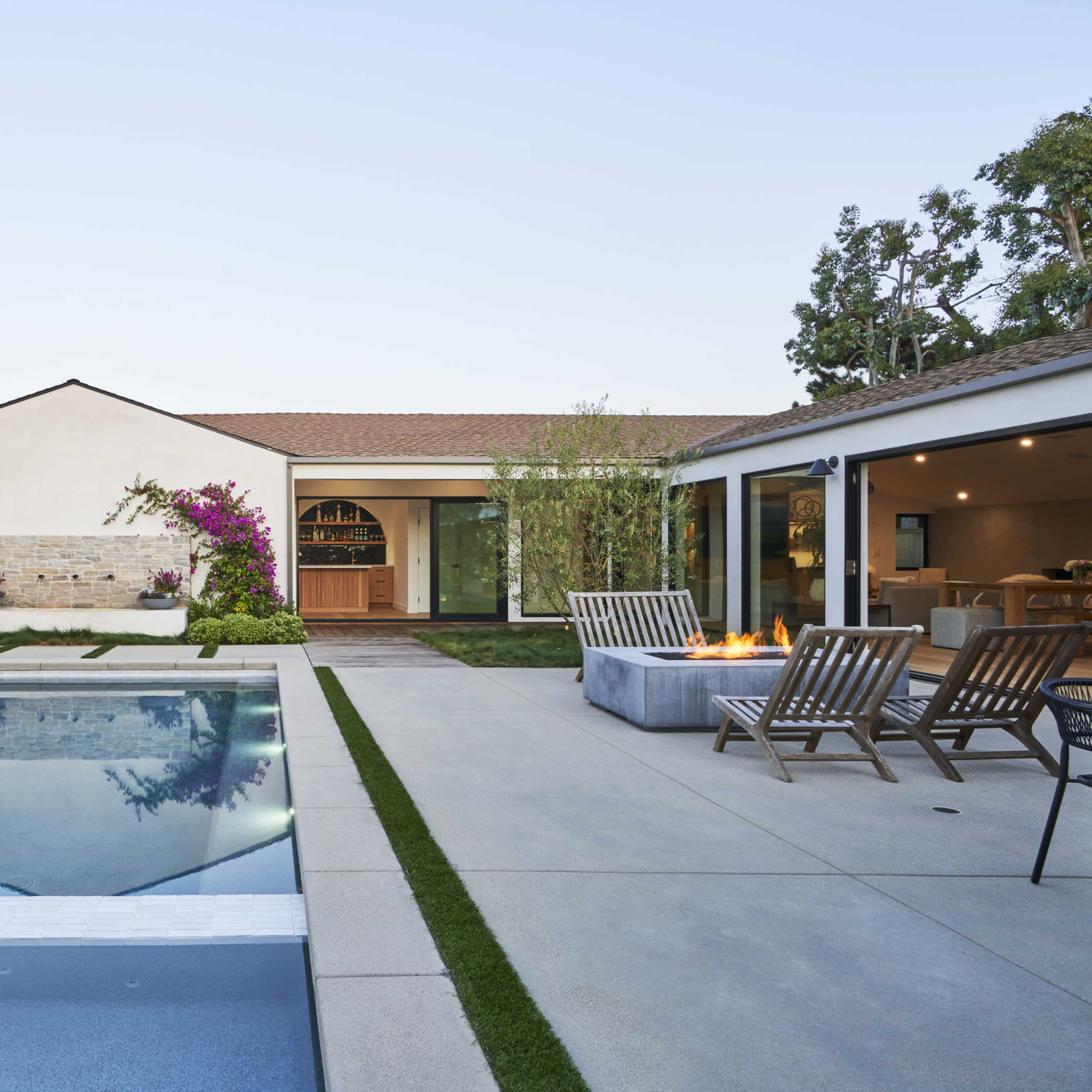 A modern backyard with a swimming pool, patio area, fire pit, and a house featuring large windows and landscaping.