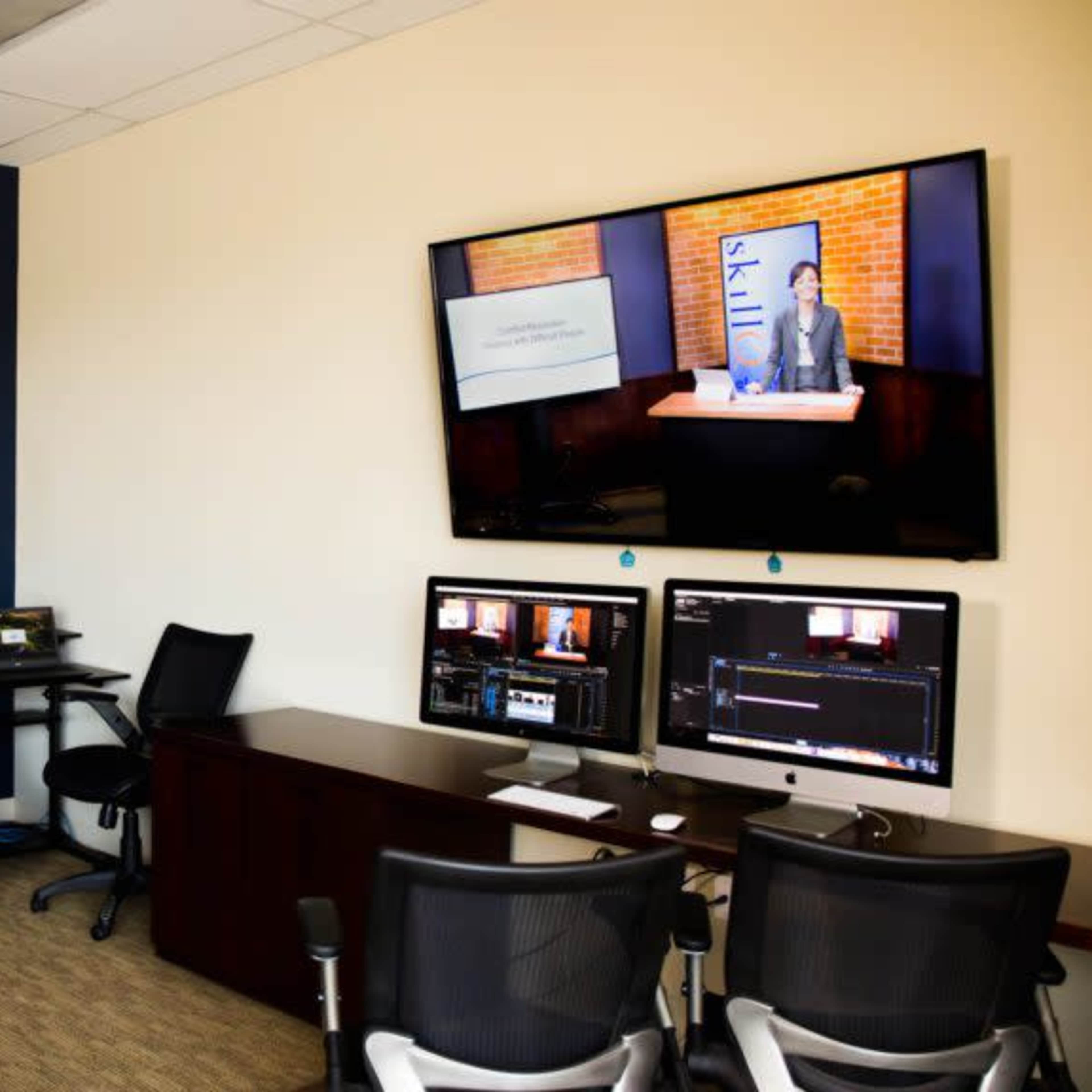 A control room features multiple screens for video production, including two large monitors on the walls and a desk with editing equipment and computers.