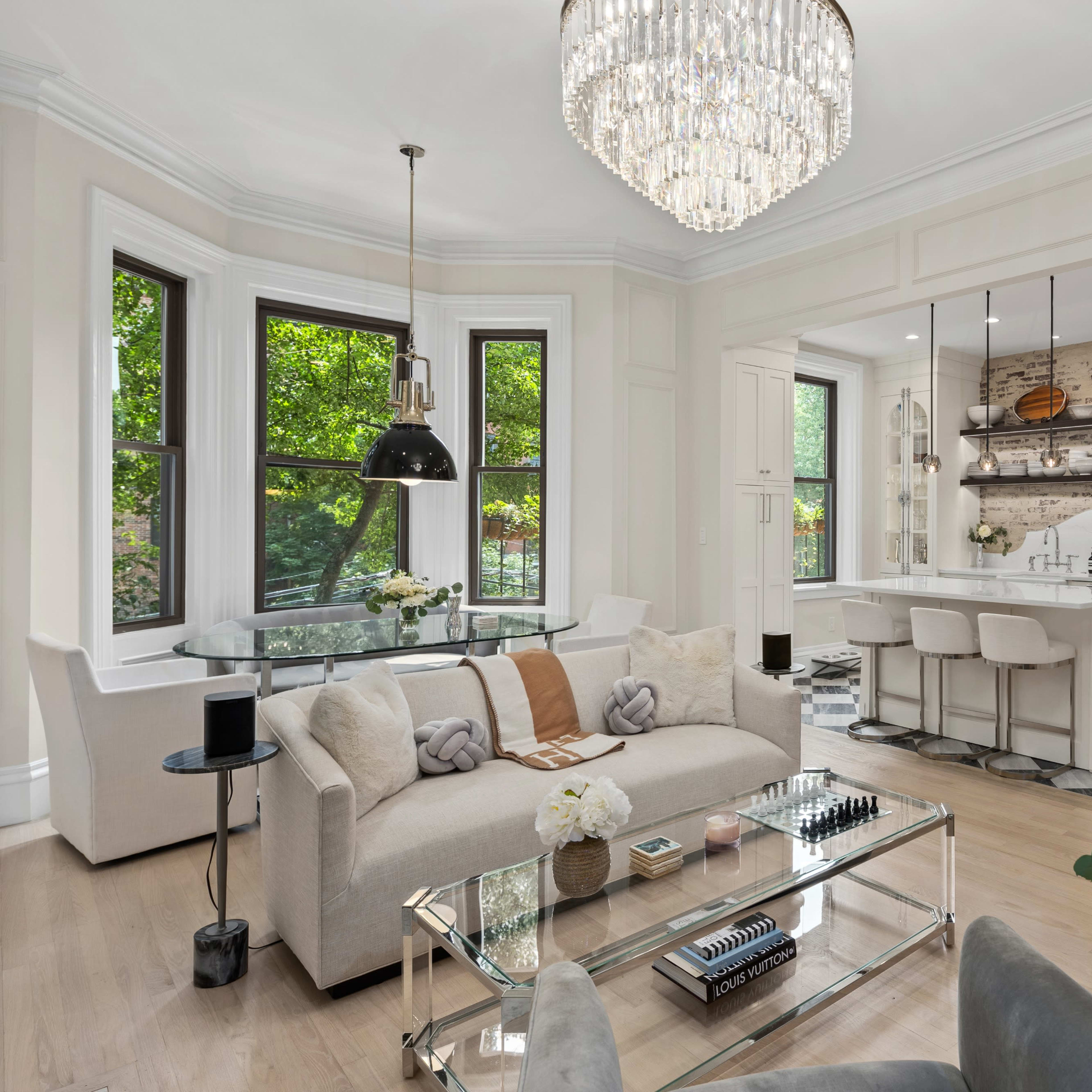 A modern living room featuring a light-colored sofa and chairs, a glass coffee table, and a kitchen visible in the background with open shelves and stylish decor.