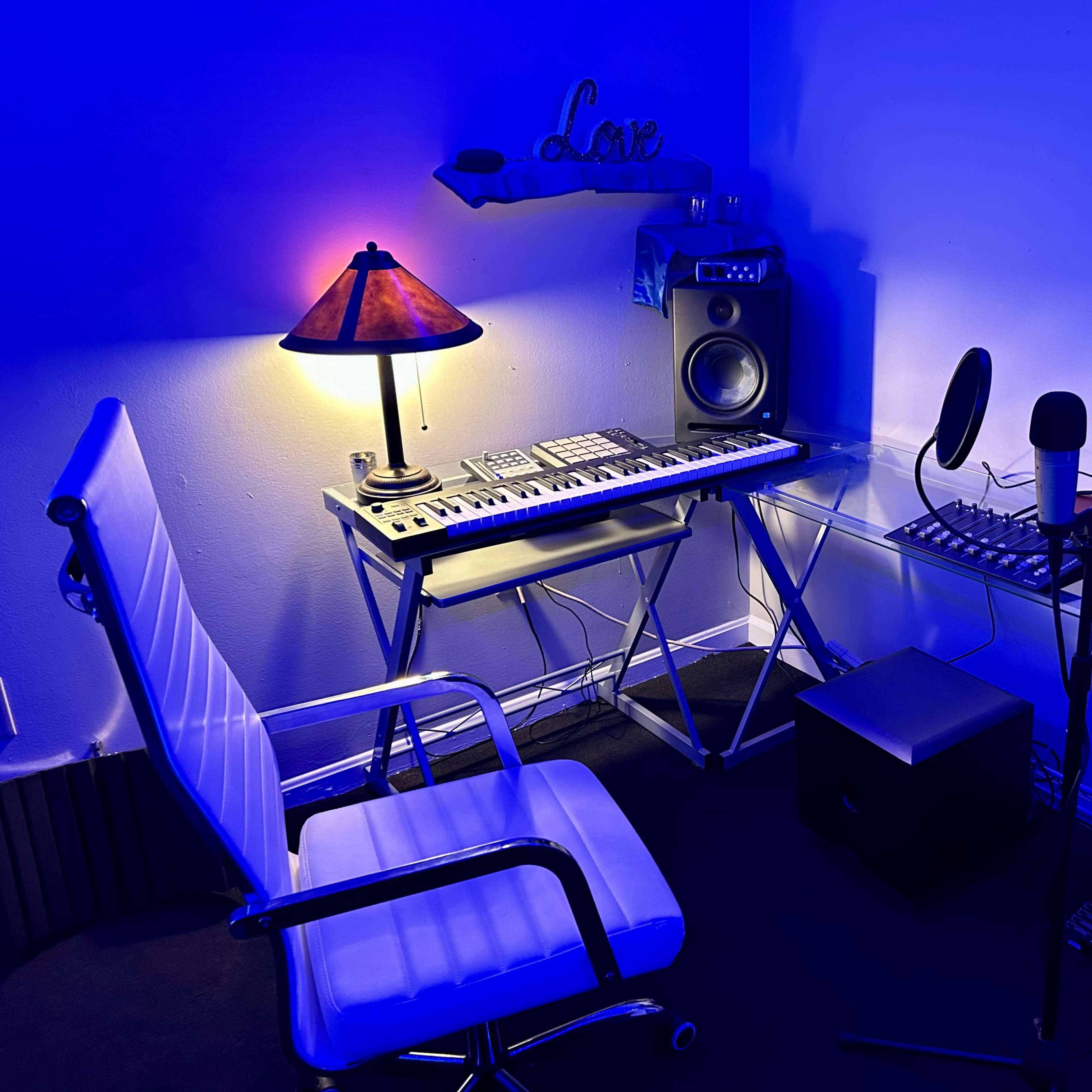 A compact music production workspace features a keyboard, studio monitors, a microphone, and a lamp illuminating a chair.