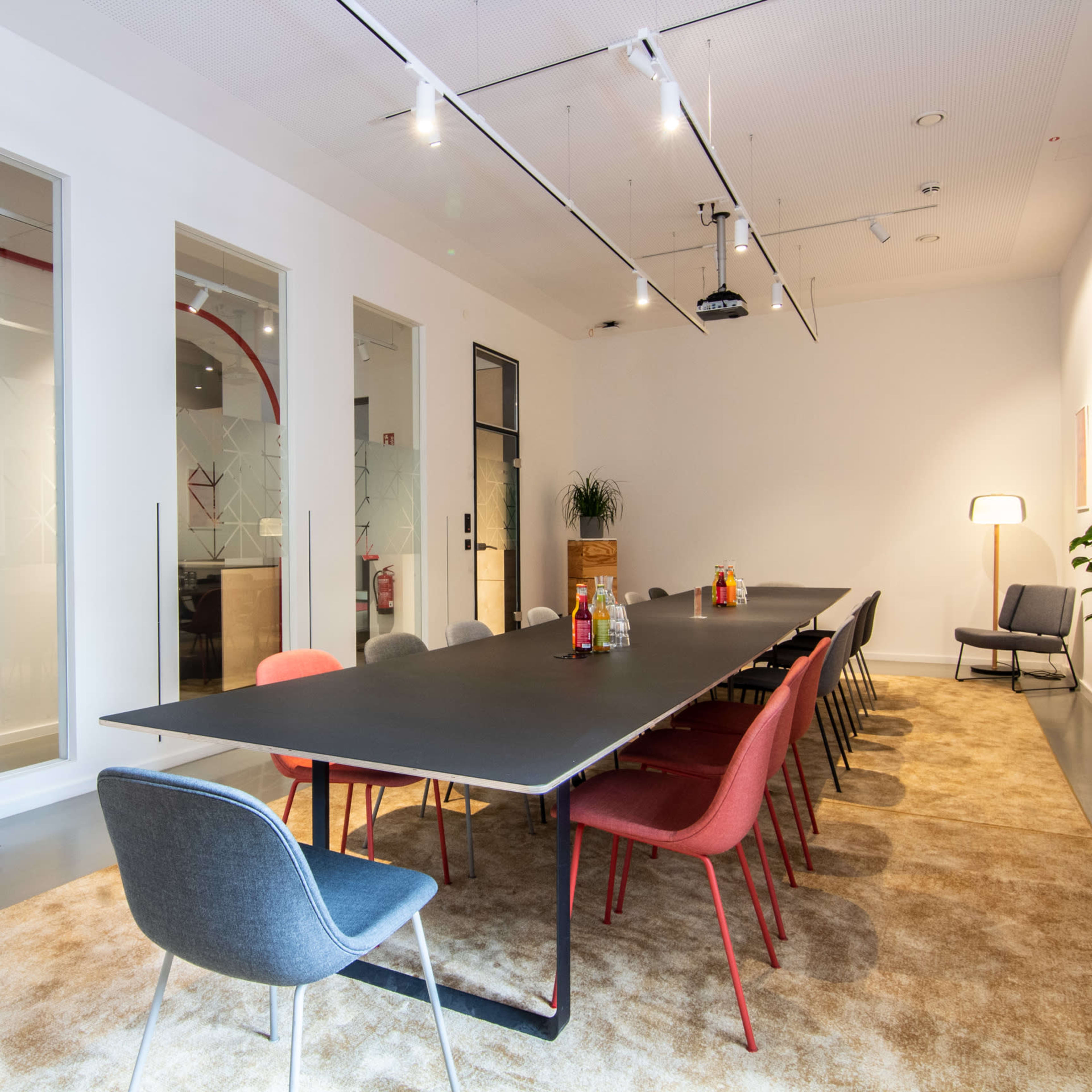 A modern conference room features a long black table surrounded by colorful chairs, with large windows allowing natural light and a carpet underfoot.