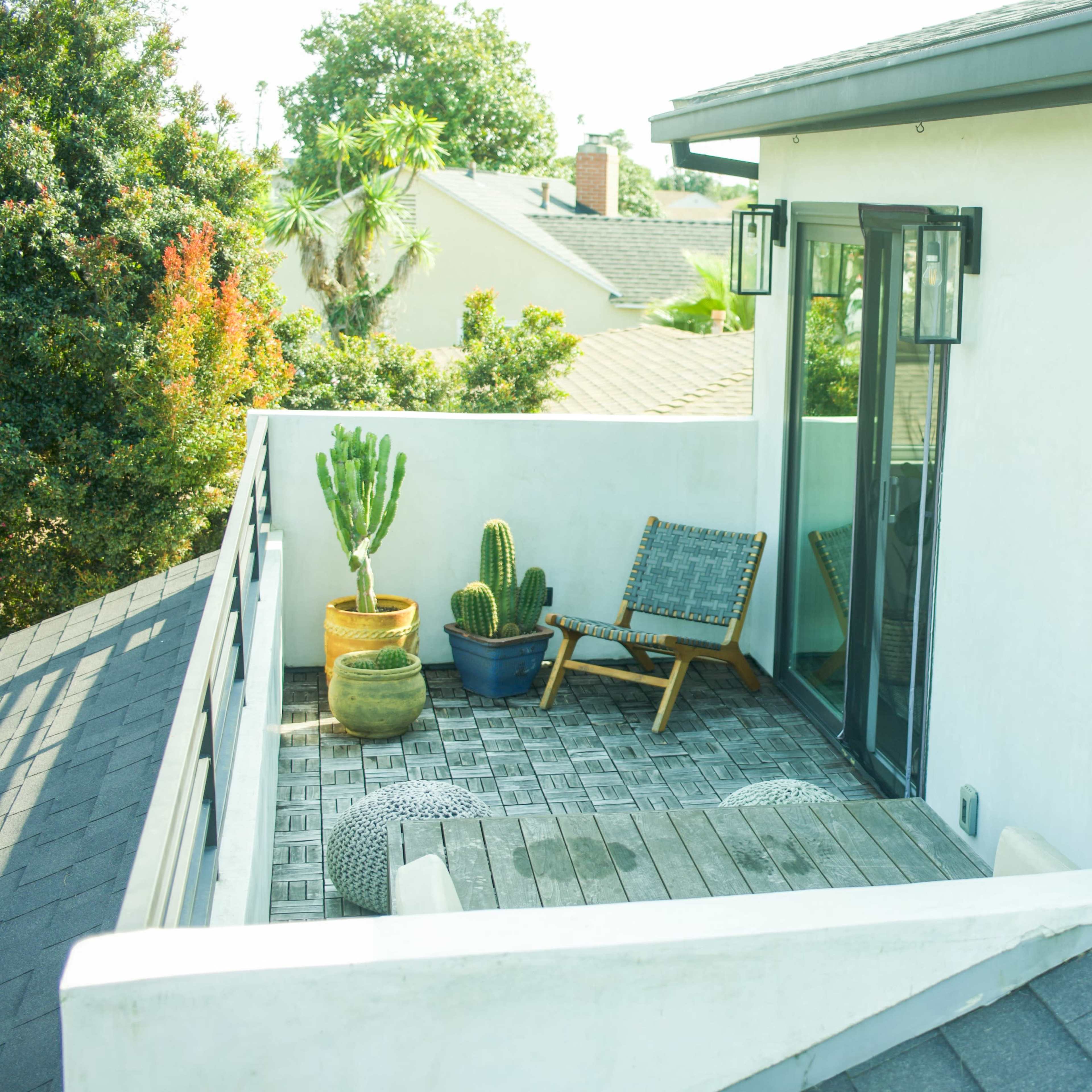 A small balcony with a wooden floor, featuring a cactus plant in a pot, a lounge chair, and a view of neighboring rooftops and greenery.