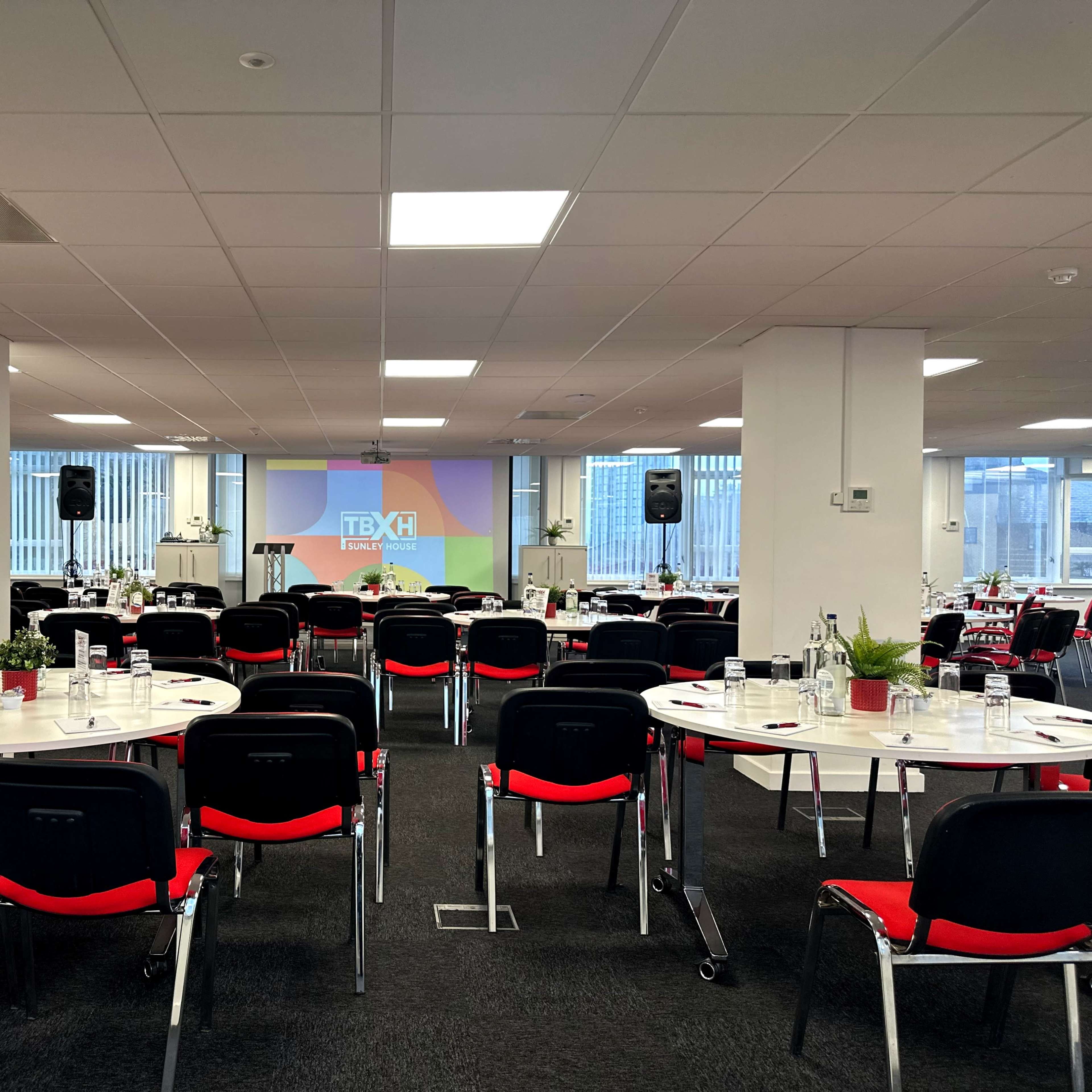 A conference room is set up with round tables arranged for a presentation, featuring chairs, plants, and a screen displaying the event logo.