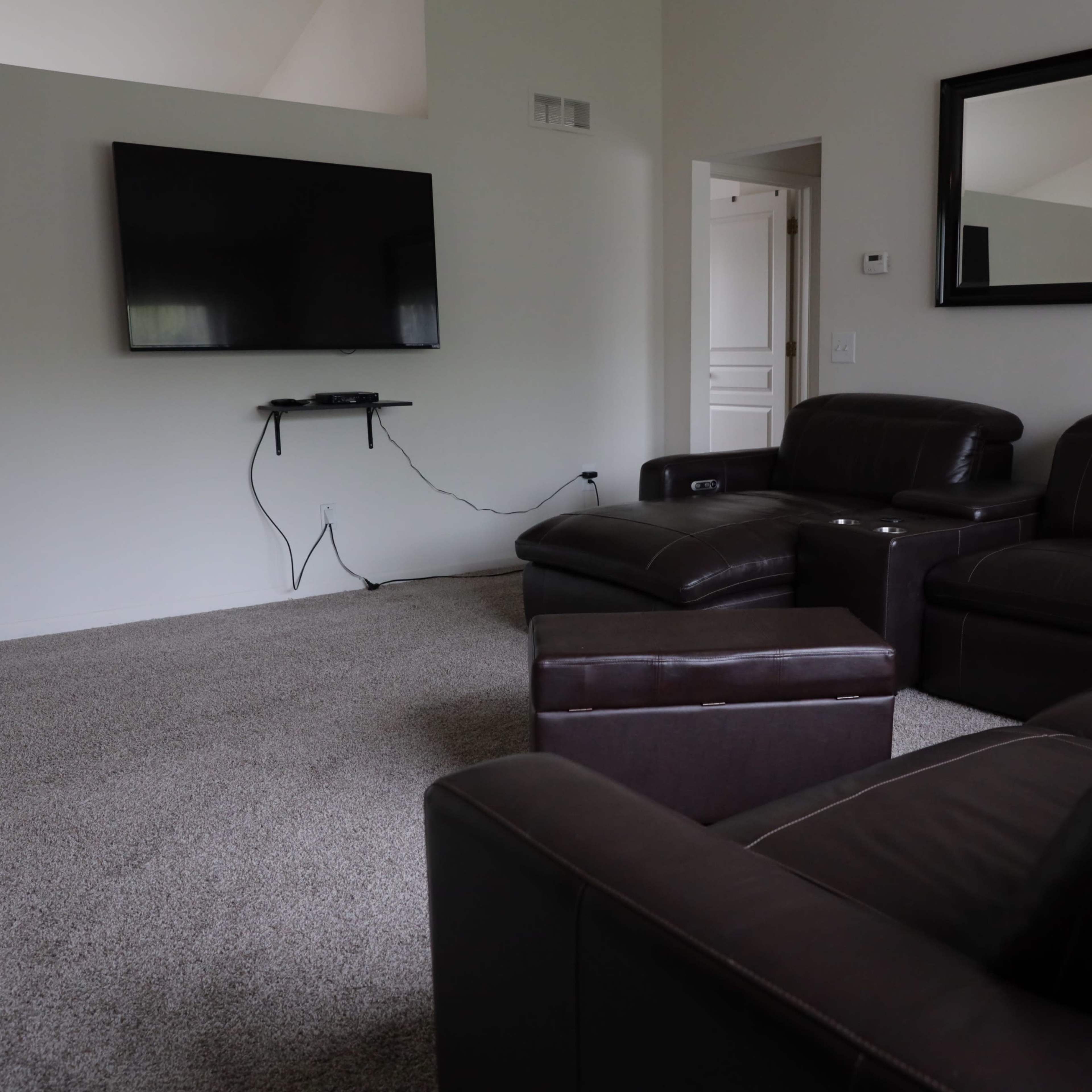 A living room with a brown sectional sofa, a large flat-screen TV mounted on the wall, and a carpeted floor.