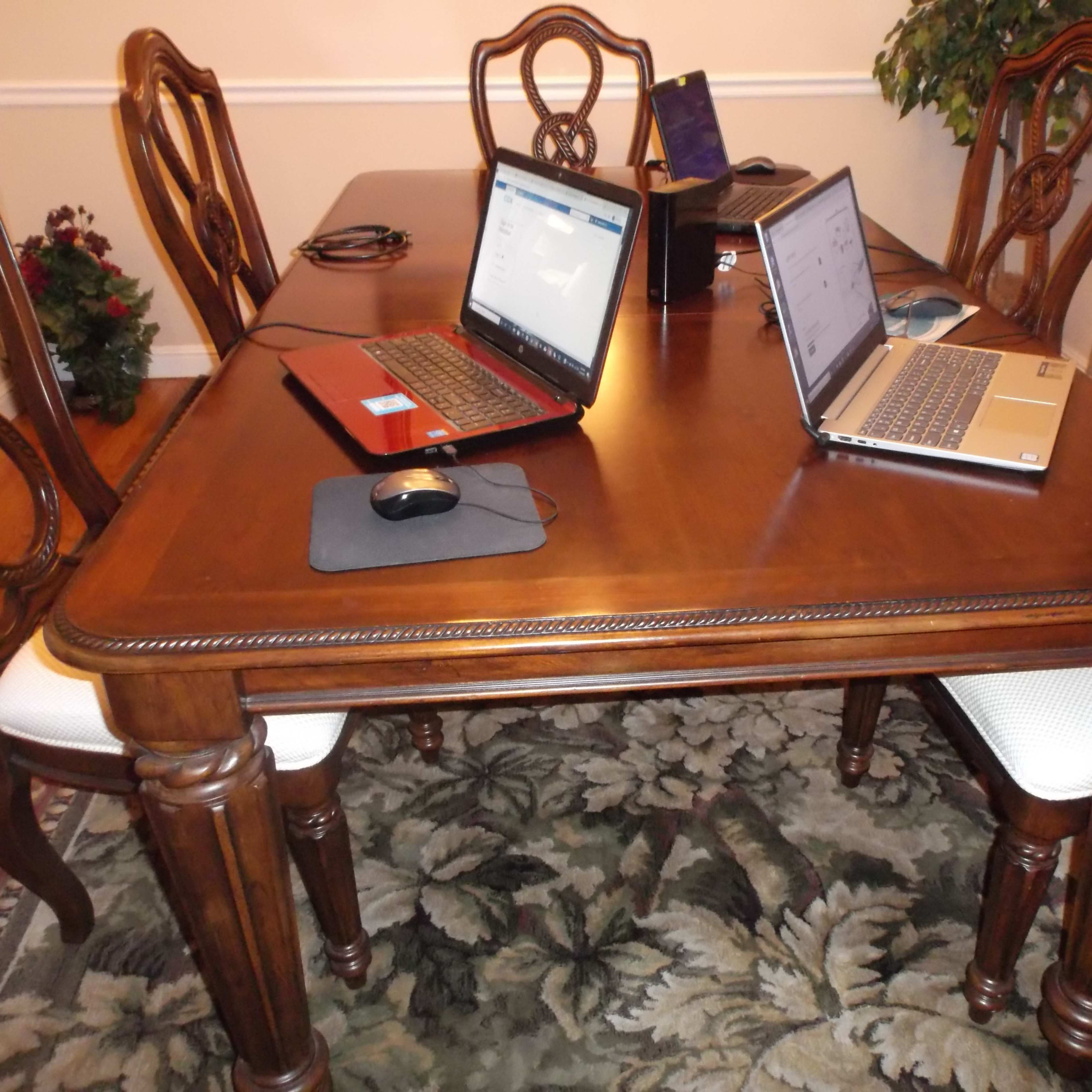 A wooden dining table is set with two laptops and a mouse, surrounded by chairs on a patterned rug.