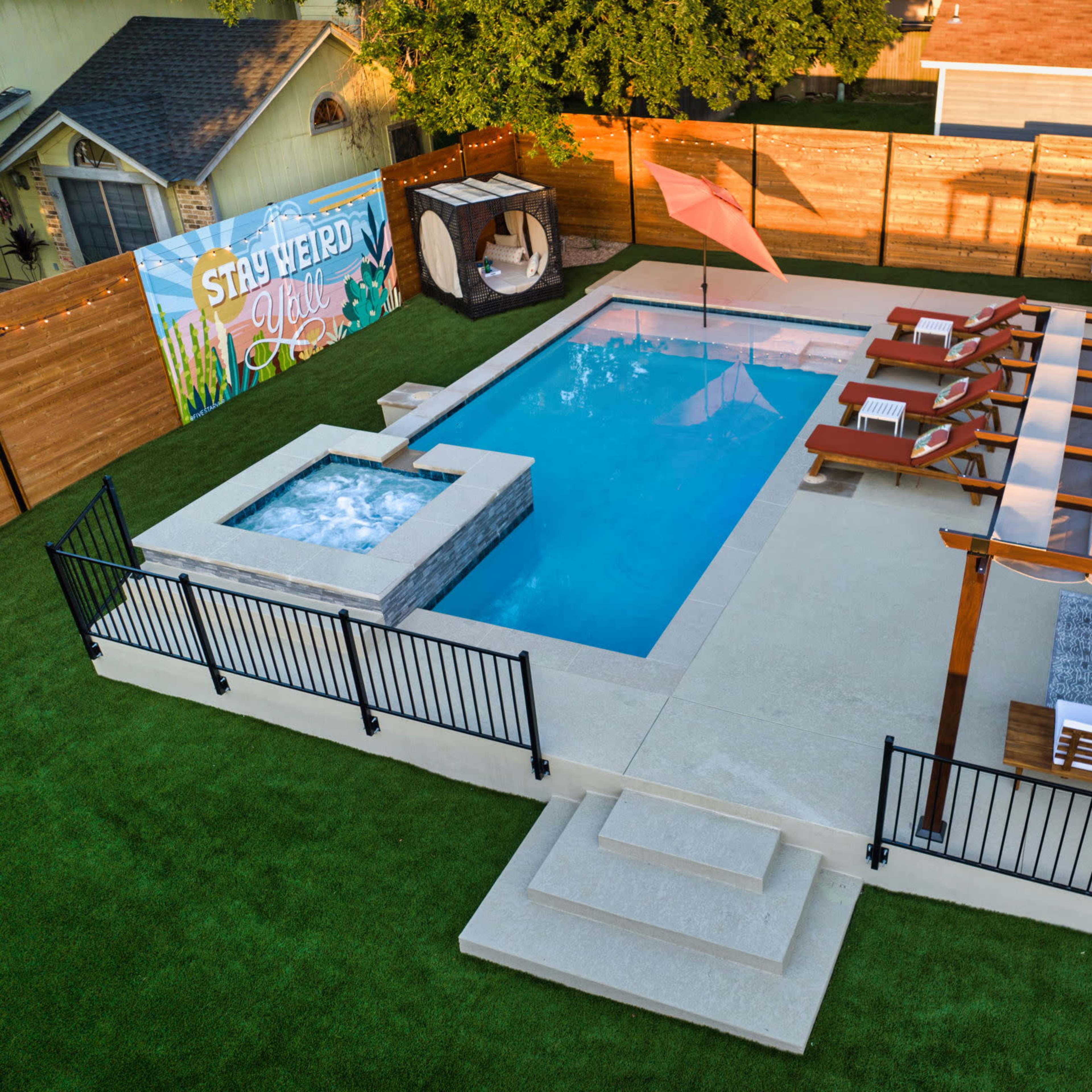 A swimming pool area with a spa, lounge chairs, and a shaded seating area surrounded by a fence and grass.