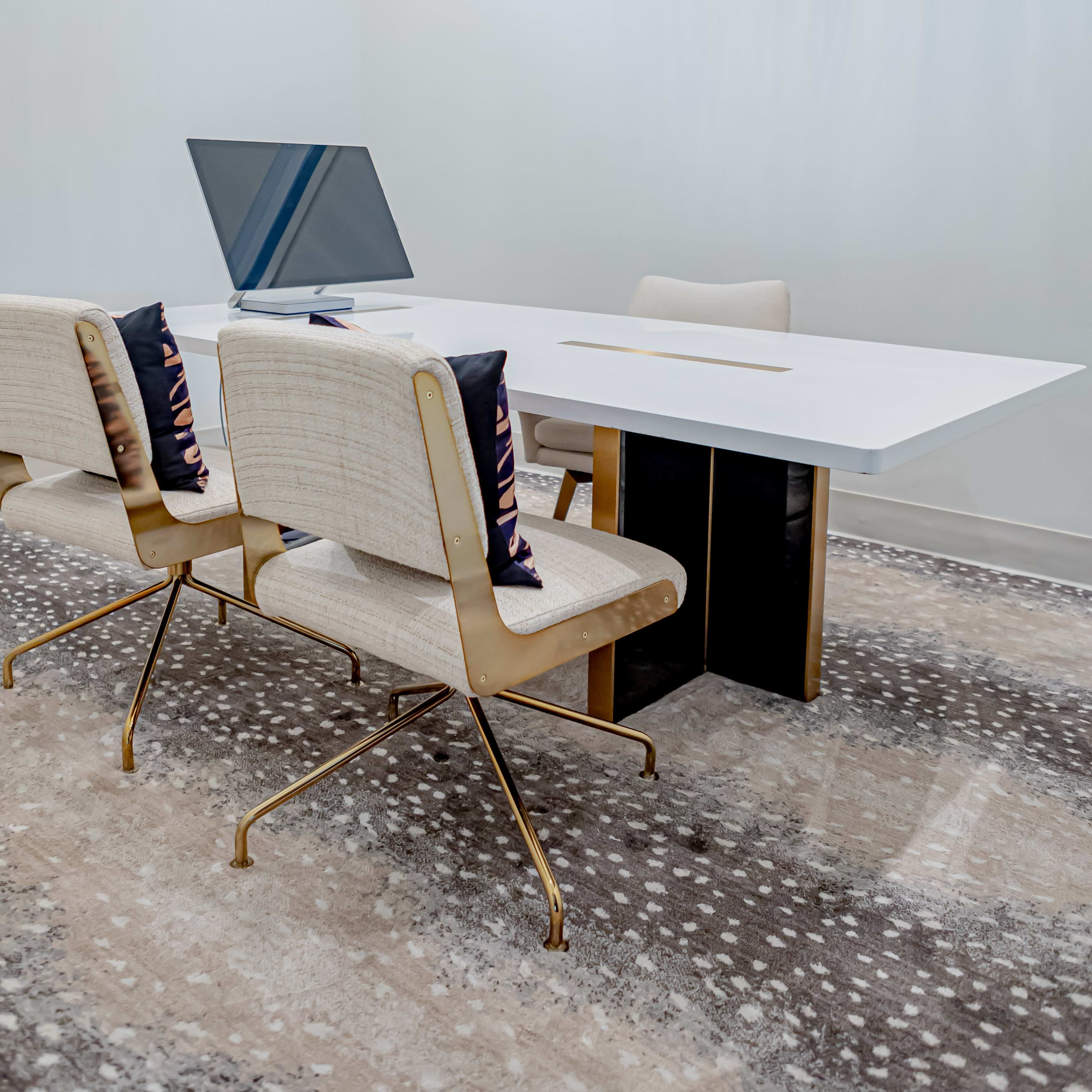A modern office space features a sleek white table, three beige chairs with cushions, and a large chandelier above, all situated on a patterned rug.